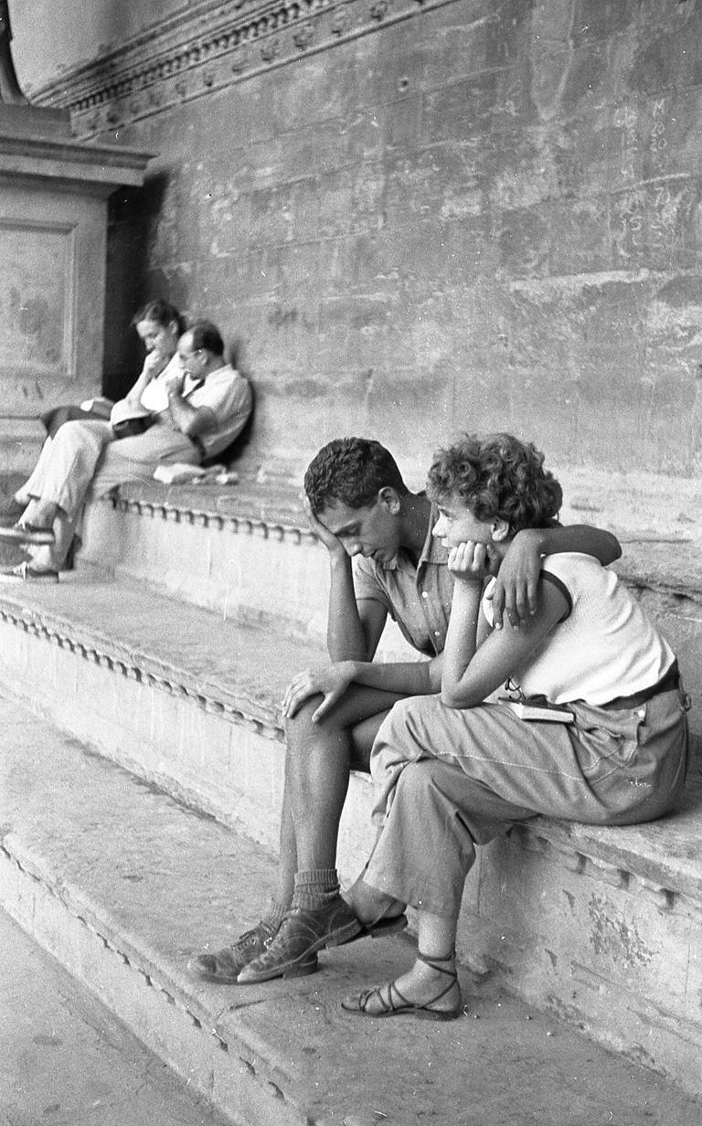 Couple on Steps in Italy von Ruth Orkin zeigt einen jungen Mann und eine Frau, die zusammensitzen und einen intimen Moment teilen. Der Mann legt seinen Arm um die Frau, und sie stützt ihren Arm auf sein Bein. Ein älteres Ehepaar sitzt in der Ferne