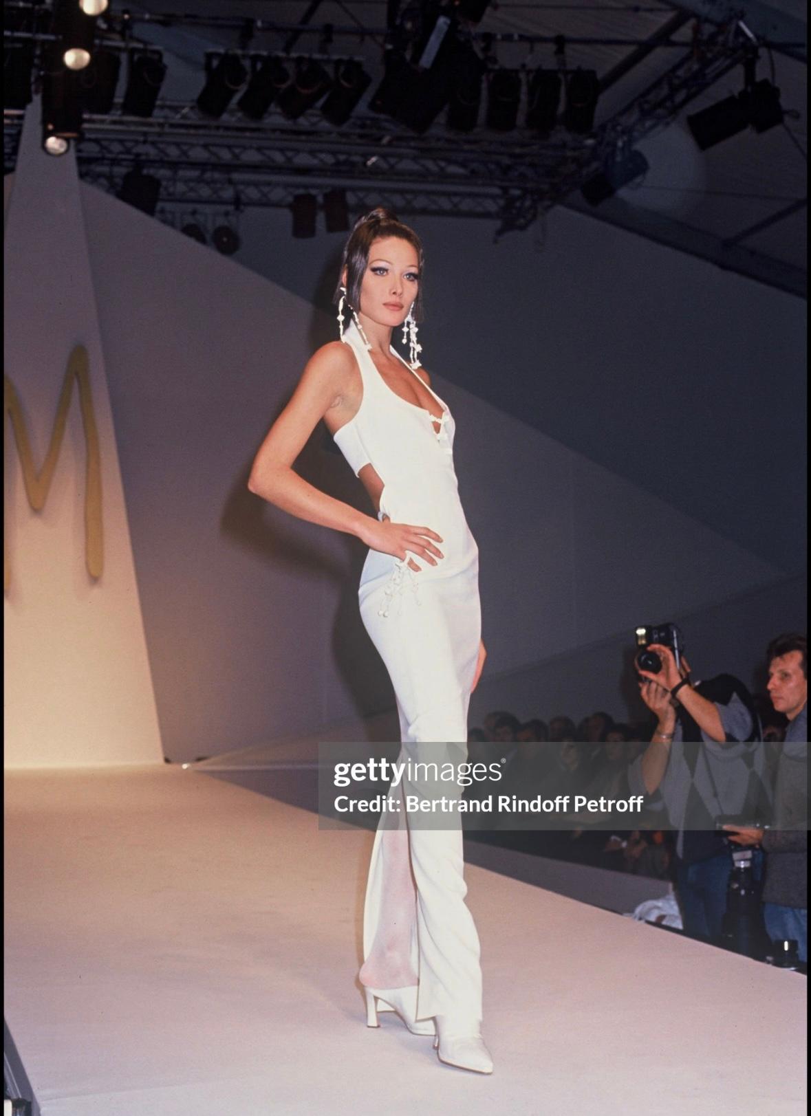 De la colección Primavera/Verano 1993 de Claude Montana, este impresionante vestido negro de cuello halter presenta un escote redondo con una pequeña caída, acentuado con cuentas blancas en el busto. Este fabuloso vestido debutó en la pasarela de la