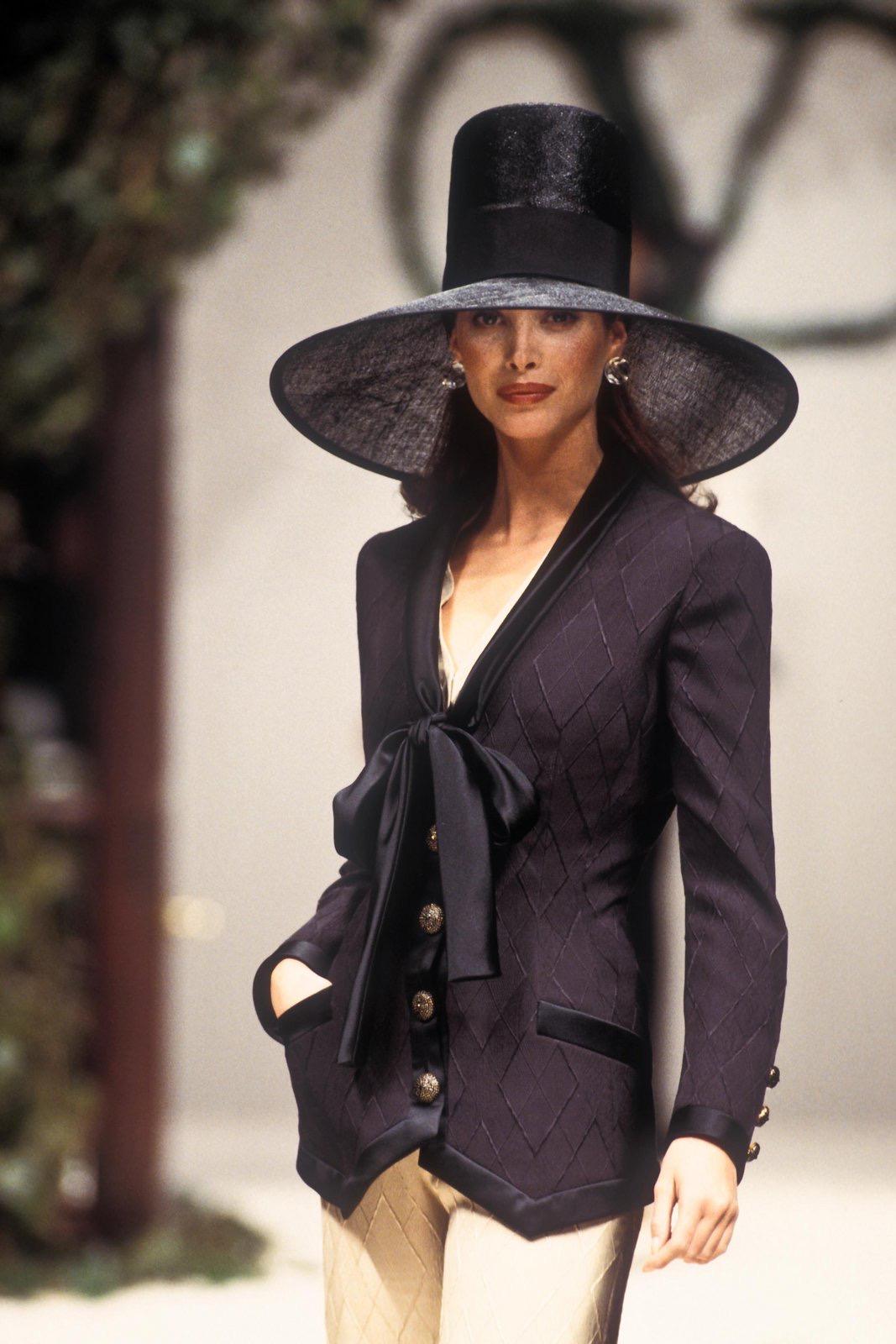 From Valentino’s Spring/Summer 1993 Haute Couture collection, this tailored black blazer jacket features a diamond-patterned jacquard fabric, satin lapels, and a dramatic silk bow tie closure. With a sharply notched hem and floral buttons, the