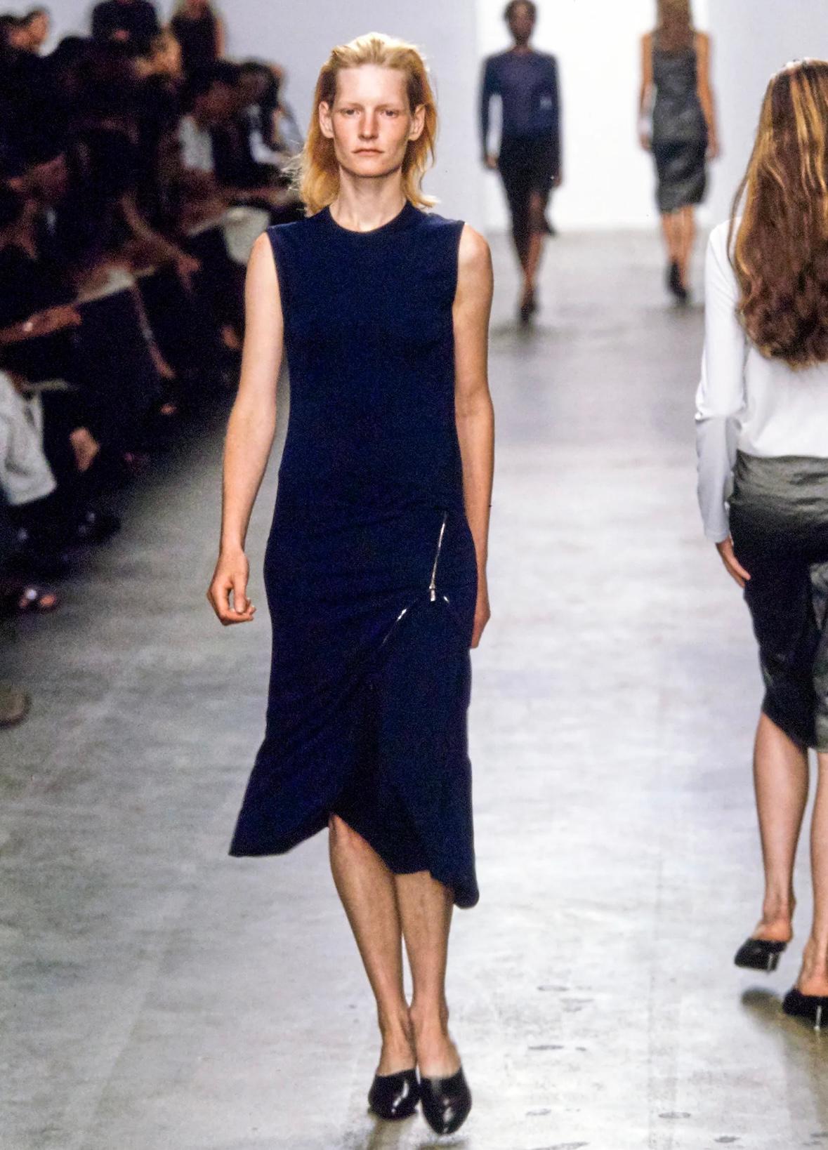 Cette robe bodycon noire sans manches de la collection printemps/été 1999 de Calvin Klein est dotée d'une fermeture éclair asymétrique. Une variante plus courte de cette robe a été présentée sur le podium dans le look 31, par Kirsten Owen. Également