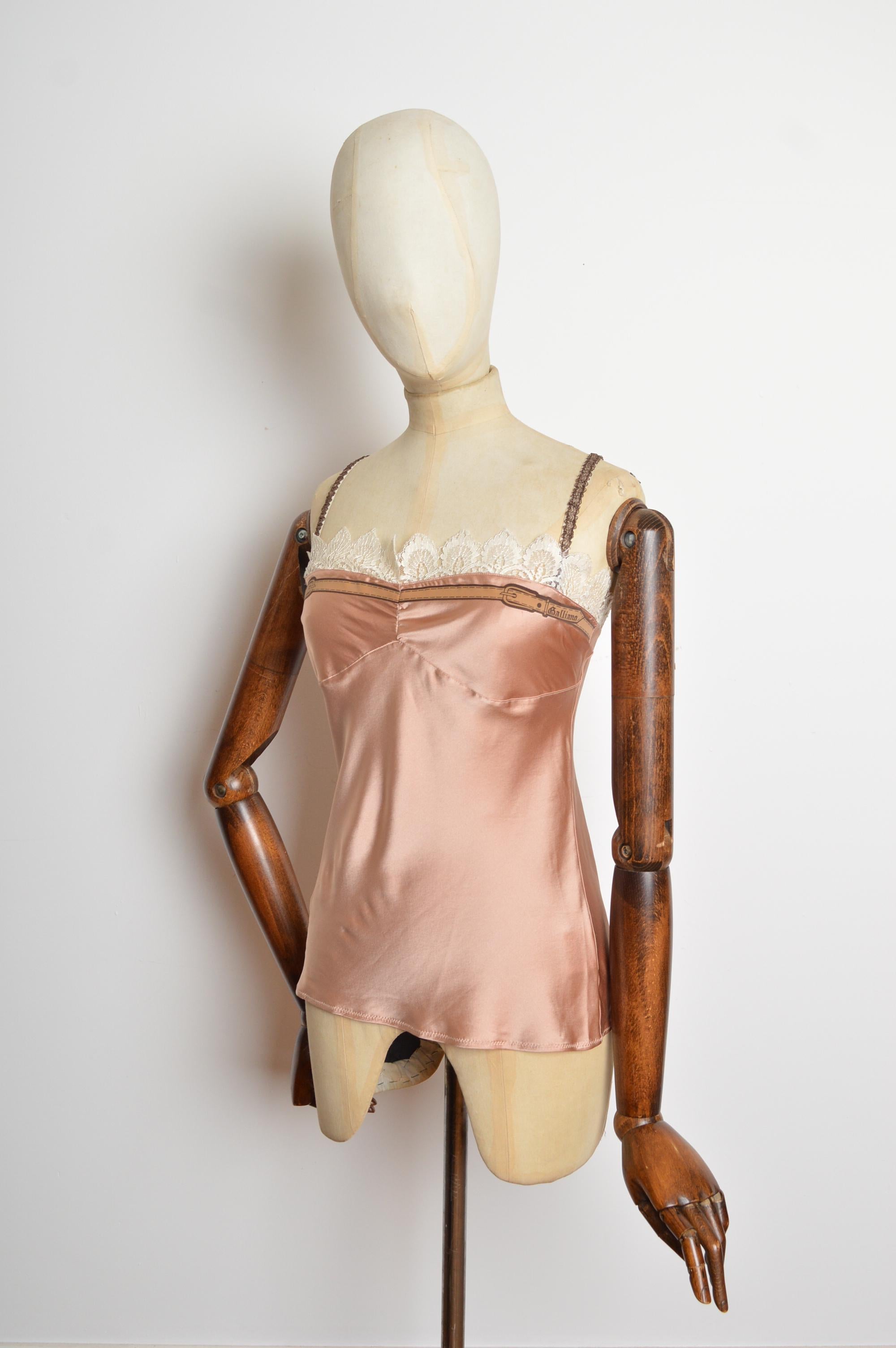 Una bellissima e delicata canotta in raso di John Galliano per la primavera/estate 2006, di colore pesca polveroso. Con cintura decorativa Galliano sul busto, finiture in pizzo Chantilly color crema e spalline delicatamente elasticizzate. 
Le misure