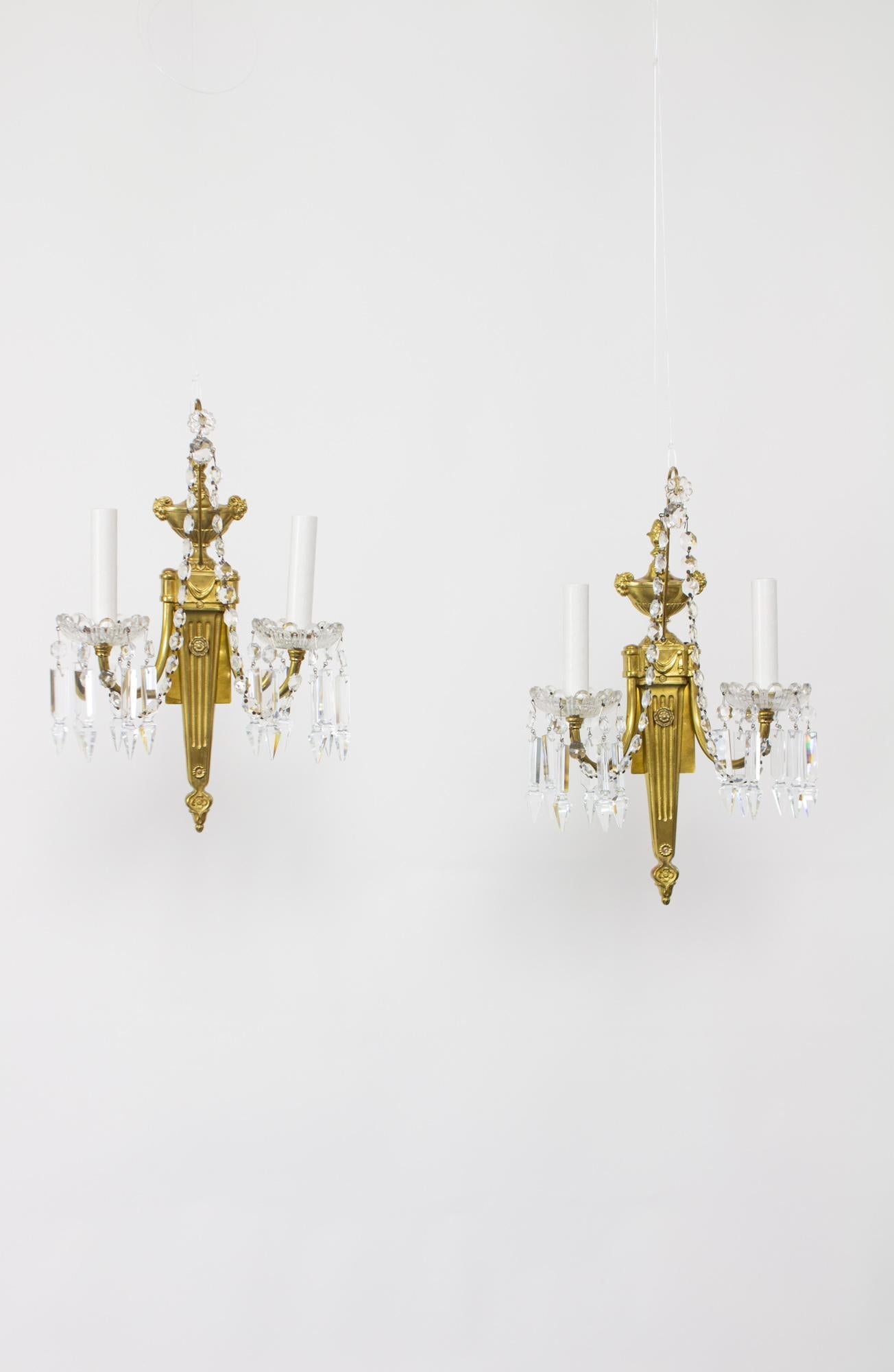 A beautiful pair of neoclassical two arm brass and crystal sconces. A long backplate with an urn form top with signature rams heads for arms. The center is column form, tapering to a rosette. The arms are simple curved brass. Crystal chains swoop