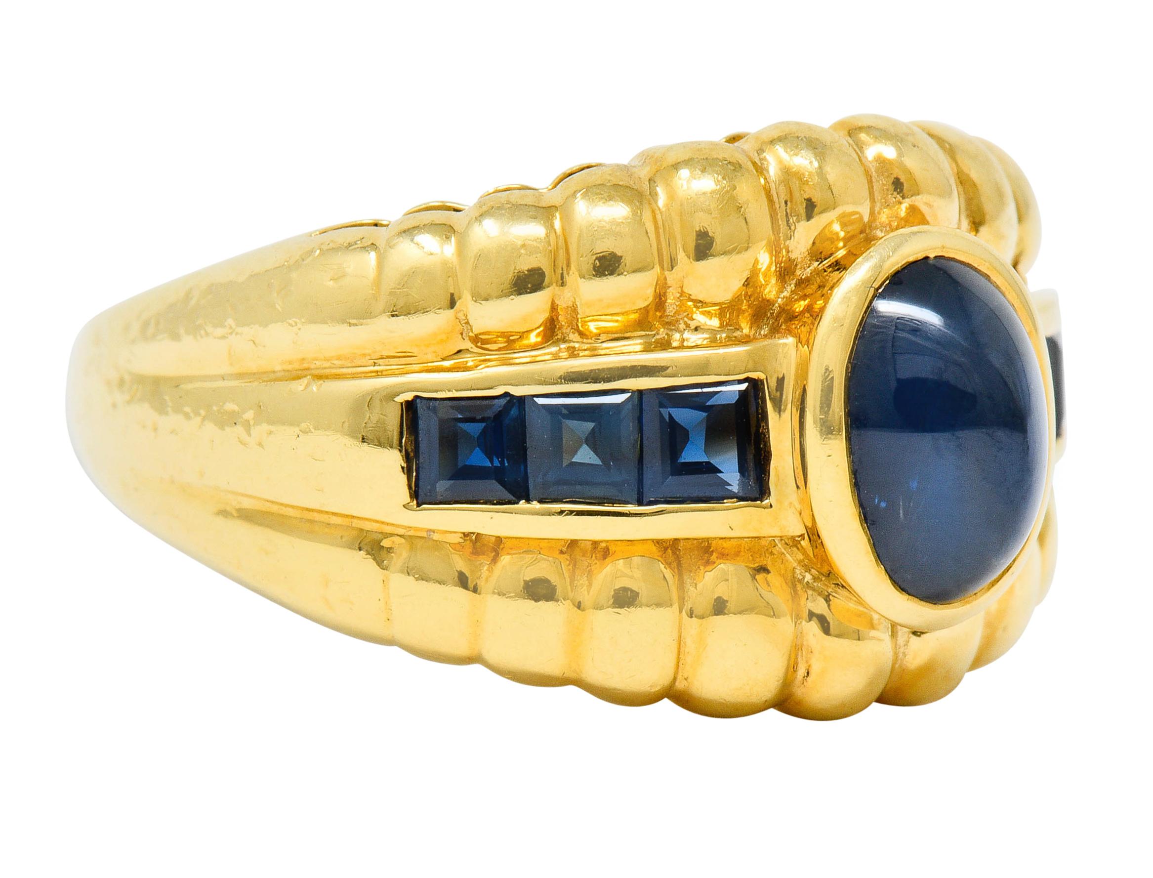 Wide band ring is brightly polished with deeply ridged edges

And centers a bezel set oval sapphire cabochon weighing approximately 1.50 carats

Translucent and medium-dark blue with subtle chatoyancy

Flanked by channel set and square cut sapphire