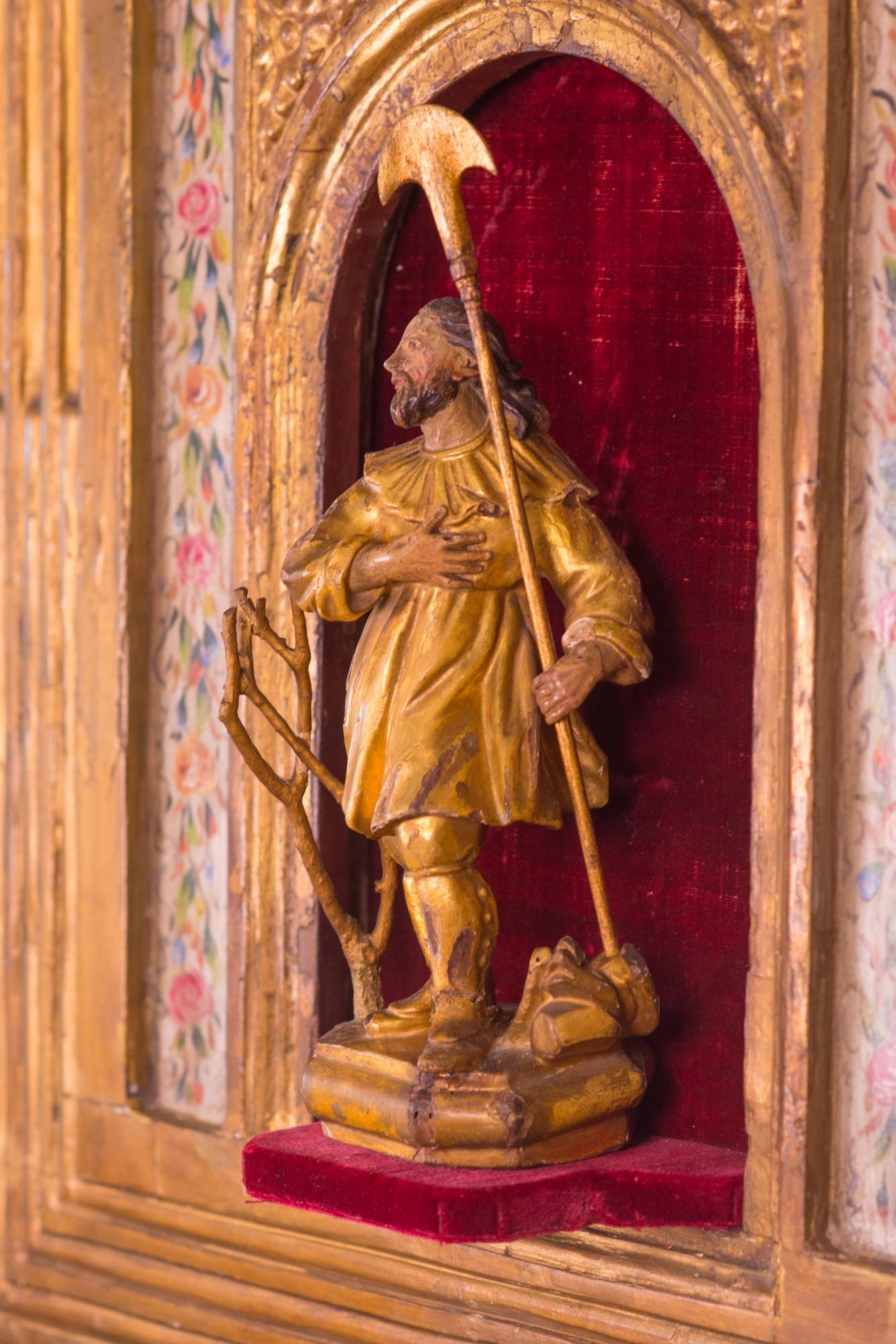 Escultura y altar españoles de madera tallada y policromada del siglo XVI de San Isidro, Patrón de la Agricultura y la Jardinería. An He está de pie en un nicho de línea de terciopelo rojo. El altar está adornado con motivos florales pintados y