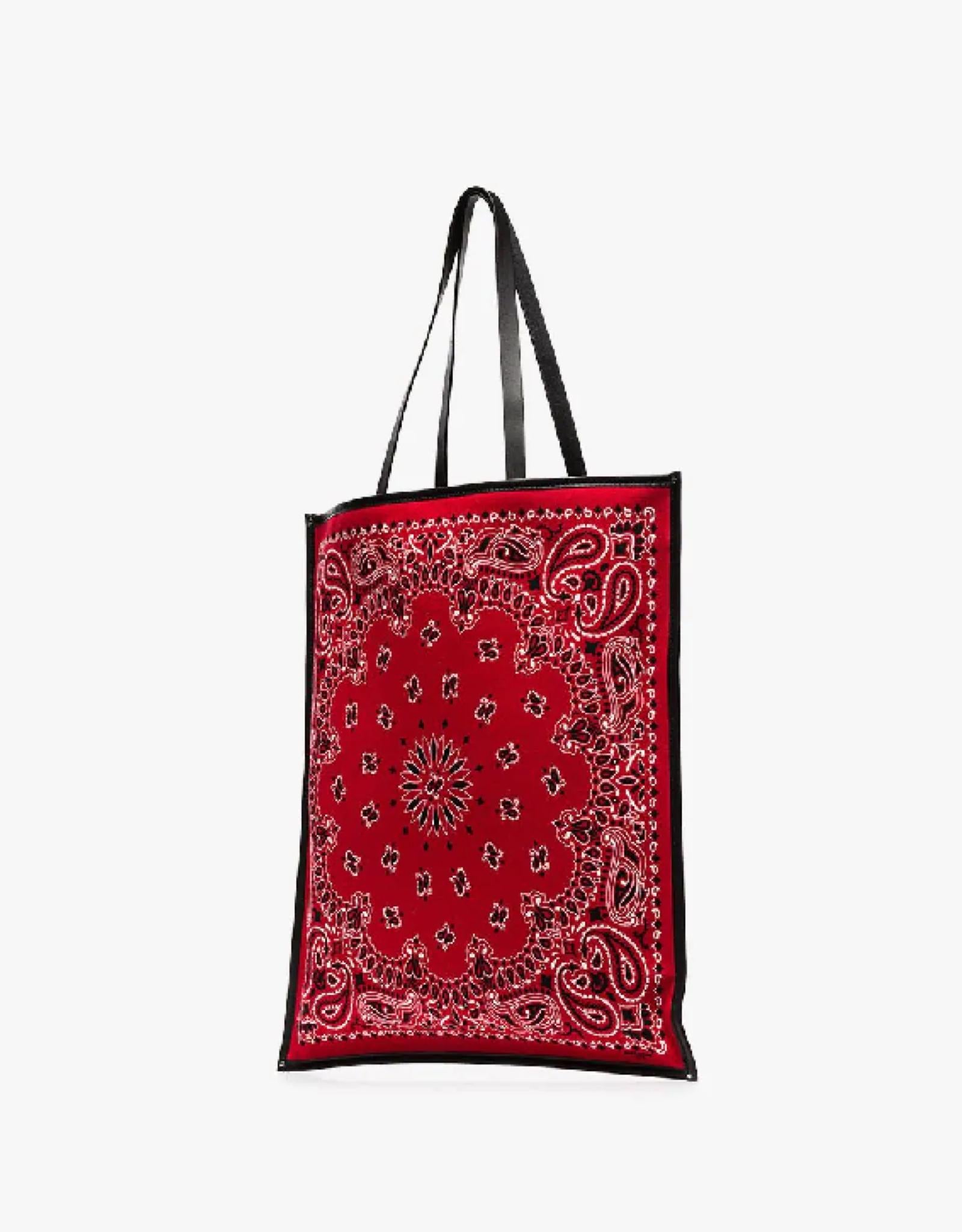 This red Saint Laurent Bandana tote bag is made from cotton and features dual flat leather top handles, a rectangular shape, leather linings and a red bandana color with a printed paisley pattern in contrasting white and black. The paisley pattern