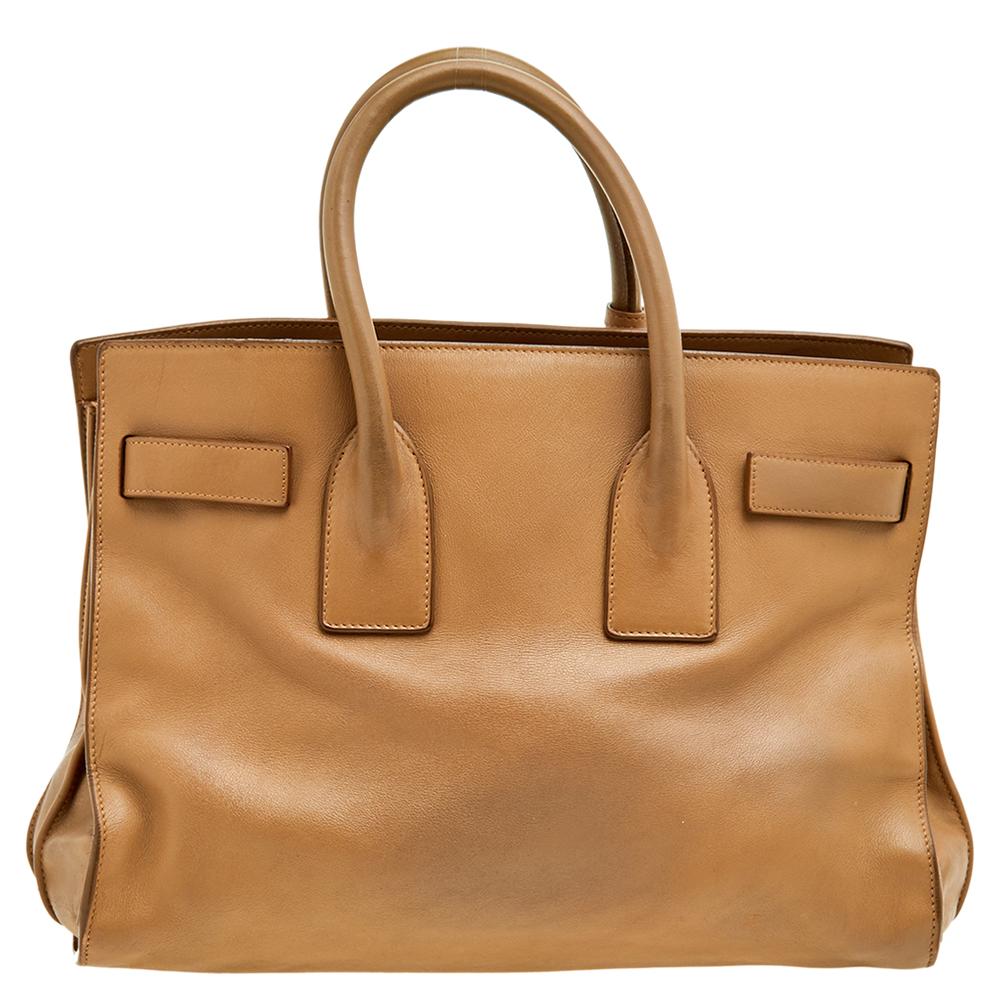 This Sac de Jour tote by Saint Laurent has a structure that simply spells sophistication. Crafted from beige leather, the bag is held by double top handles. The tote comes with a suede-lined interior with enough space to store your necessities and