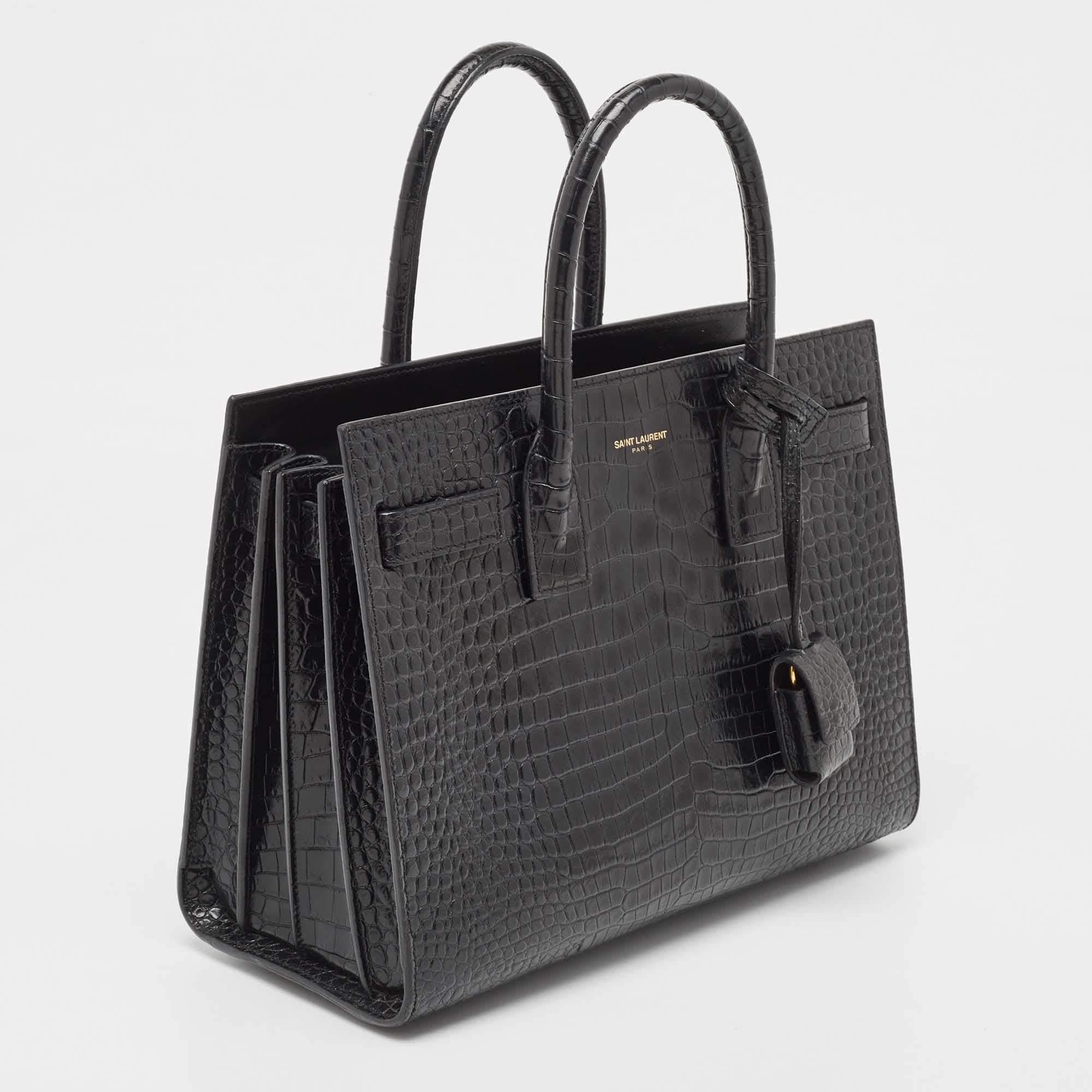 Ce sac à main Sac de Jour de Saint Laurent a une structure qui est tout simplement synonyme de sophistication. Confectionné en cuir noir gaufré au crocodile, ce sac est maintenu par deux anses supérieures. Le fourre-tout est doté d'un intérieur
