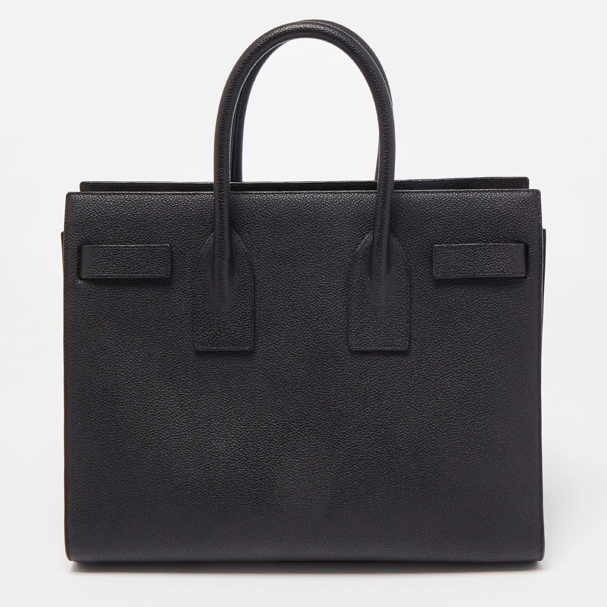 The Saint Laurent Classic Sac De Jour tote is an elegant and timeless handbag. Crafted from high-quality grain leather, it features a sleek black color, top handles, a removable shoulder strap, and the brand's signature accordion sides. The interior