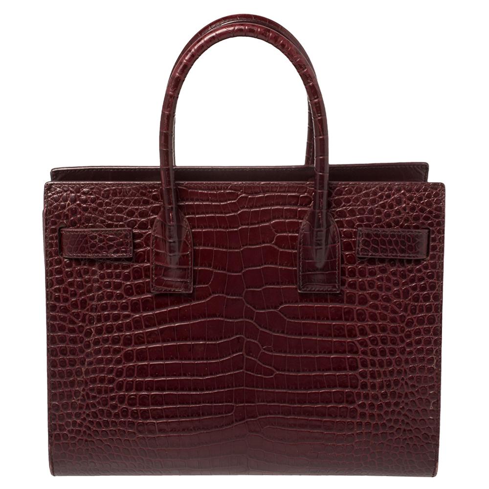 This Sac de Jour tote by Saint Laurent has a structure that simply spells sophistication. Crafted from croc-embossed leather in a burgundy shade, the bag is held by double top handles. The tote comes with a leather-lined interior with enough space