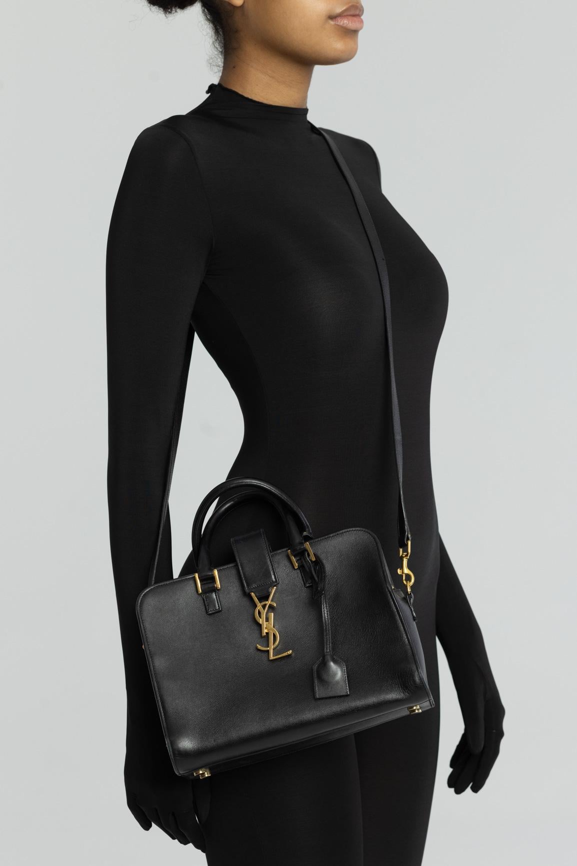 This bag is made of smooth calfskin leather in black. The bag features rolled leather top handles and a crossover strap with a weighted gold YSL monogram. The top zippers open to a matching black calfskin leather interior with zipper and patch