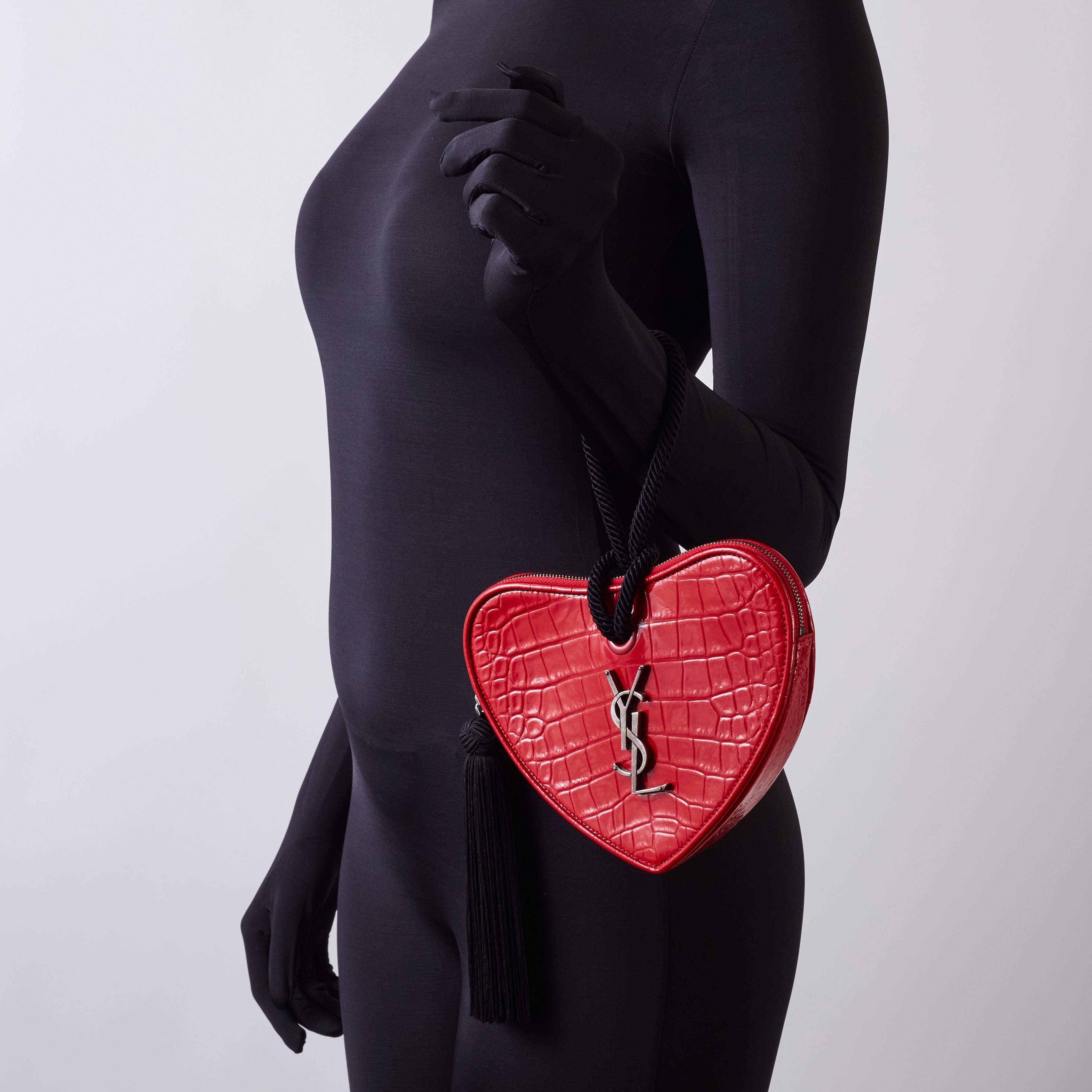 Cette pochette est réalisée en cuir rouge gaufré en forme de cœur. Sac coeur est un sac français. Le sac à main est orné d'un logo ysl en argent vieilli, d'une poignée poignet en corde noire et d'une tirette de fermeture à gland noire. La fermeture