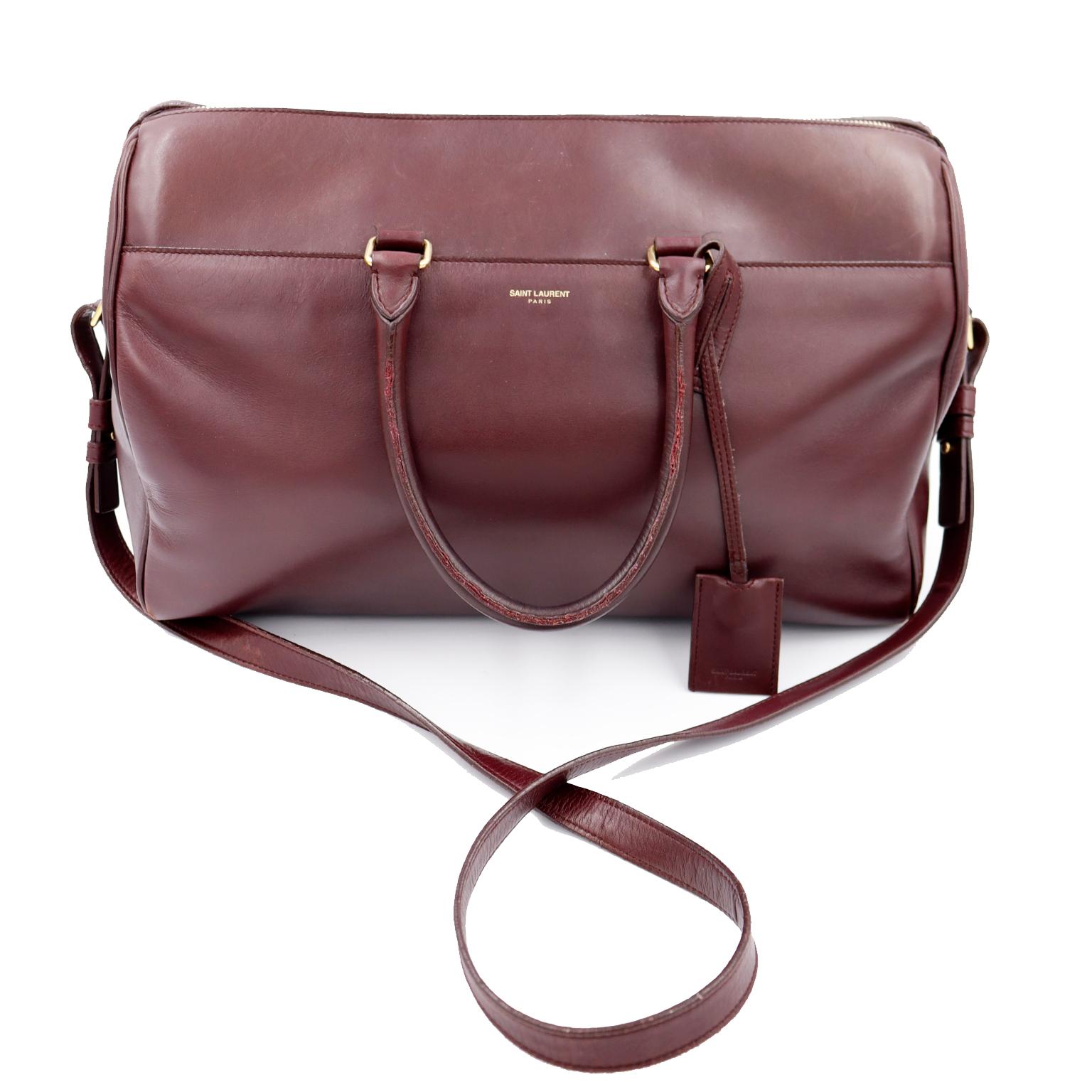 This deep burgundy leather Saint Laurent large duffle bag has double handles, a top zipper and a removable shoulder strap. The bag has brass hardware, including brass feet and a brass branded zipper pull. There is an outside open pocket and an