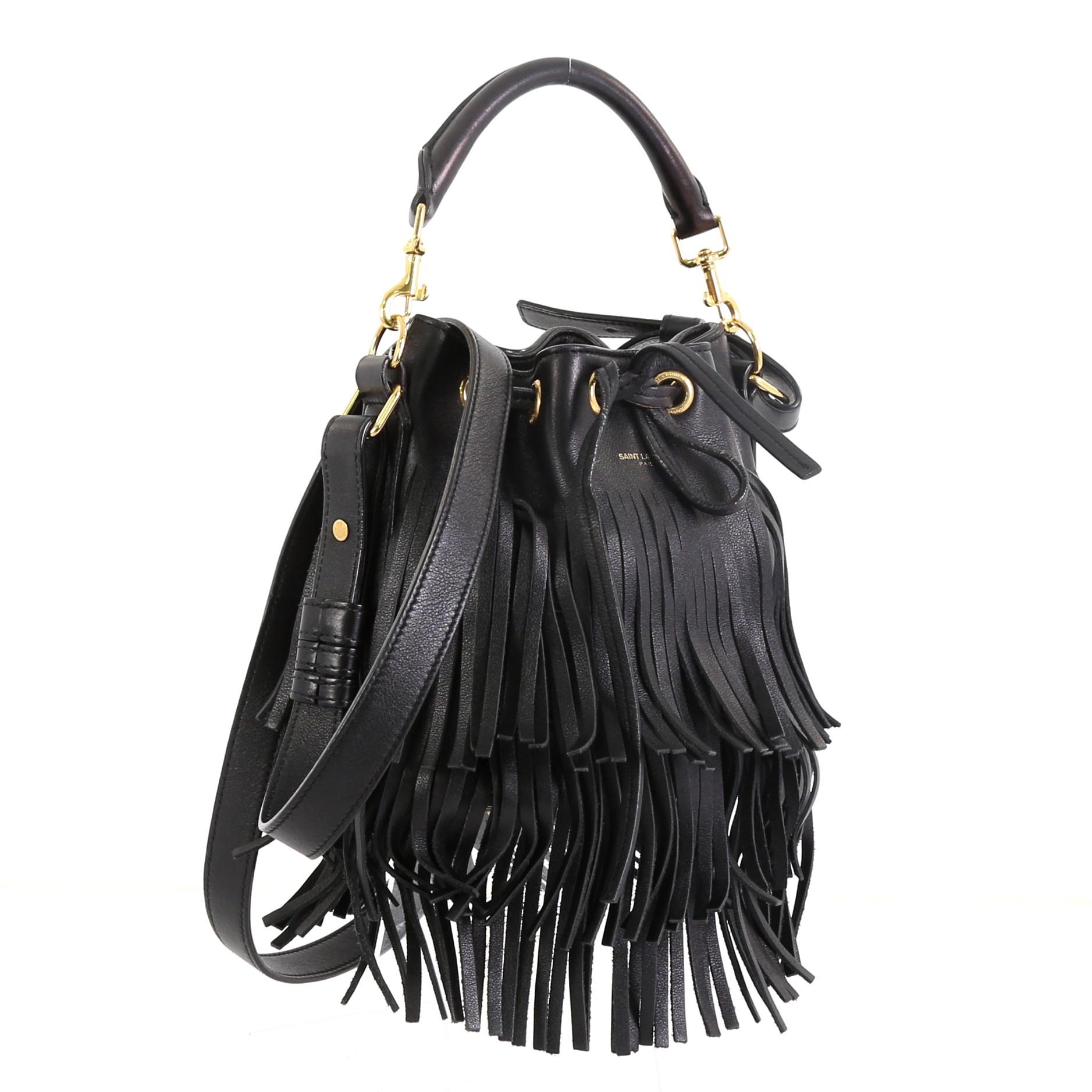 This Saint Laurent Fringe Emmanuelle Bucket Bag Leather Small, crafted in black leather, features a detachable looped handle, adjustable strap, cascading long fringes, side zip pockets, and gold-tone hardware. Its drawstring closure opens to a black