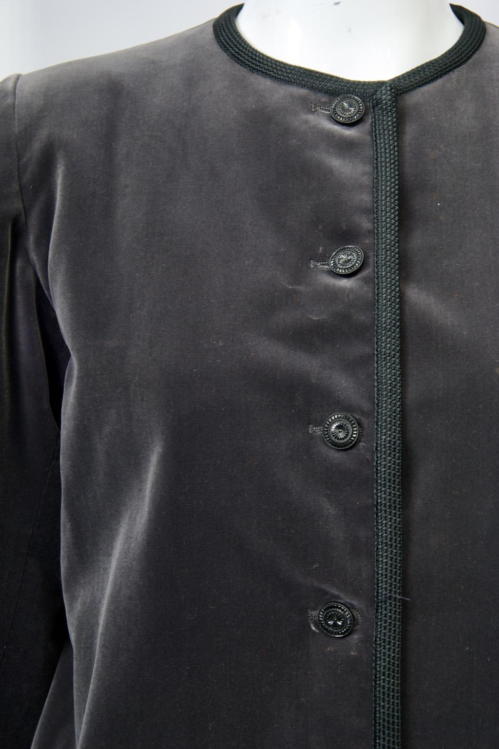 Yves Saint Laurent Rive Gauche-Jacke aus anthrazitfarbenem Samt, ca. Ende der 1970er-/Anfang der 1980er-Jahre, an allen Rändern mit schwarzer Borte eingefasst. Die kragenlose Jacke ist kurz geschnitten, hat schmale Ärmel und fünf Knöpfe im Jet-Stil.