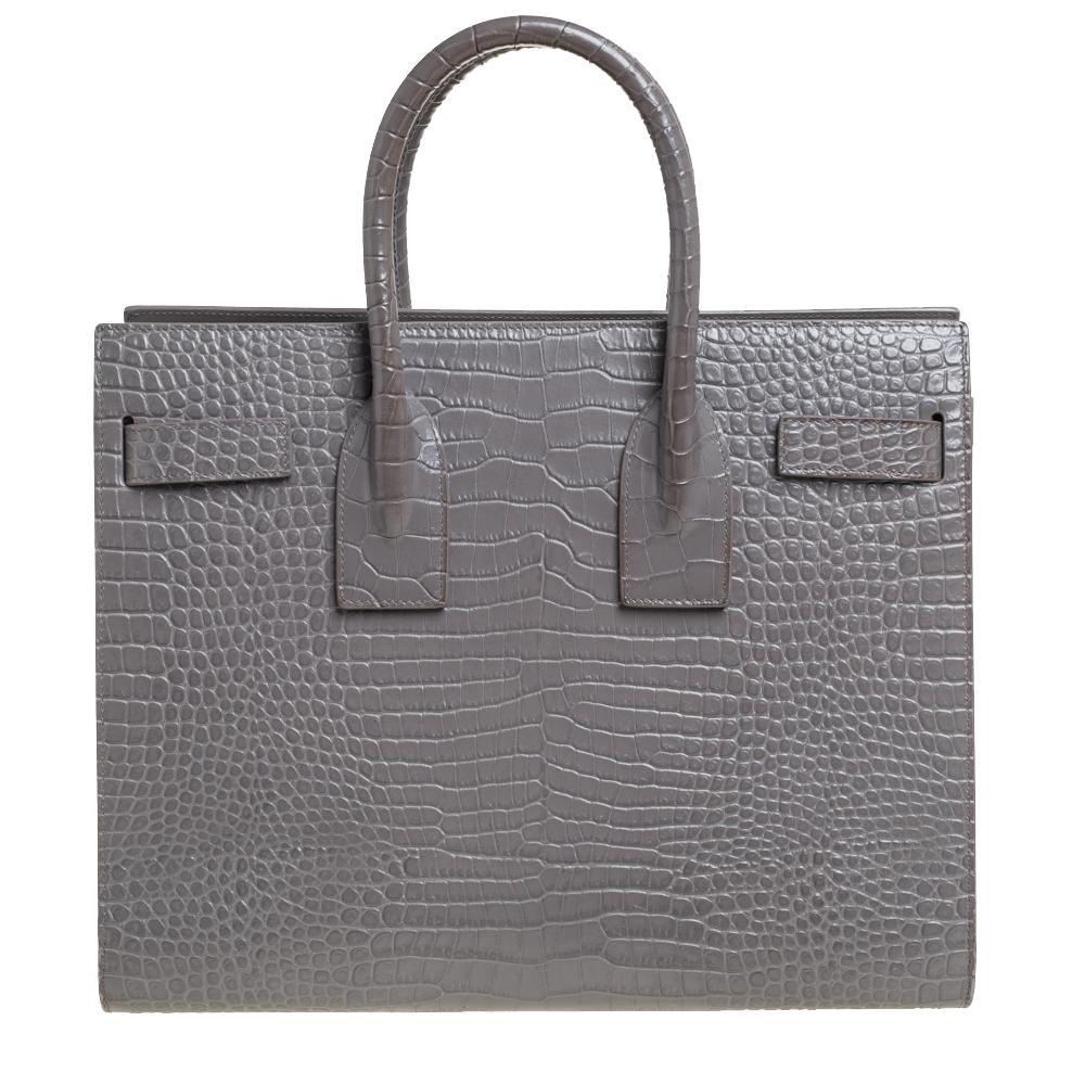 This Sac de Jour tote by Saint Laurent has a structure that exhibits a sophisticated image. Crafted from grey croc-embossed leather, the bag is held by double top handles and a shoulder strap. The tote comes with a lined interior and the bottom is