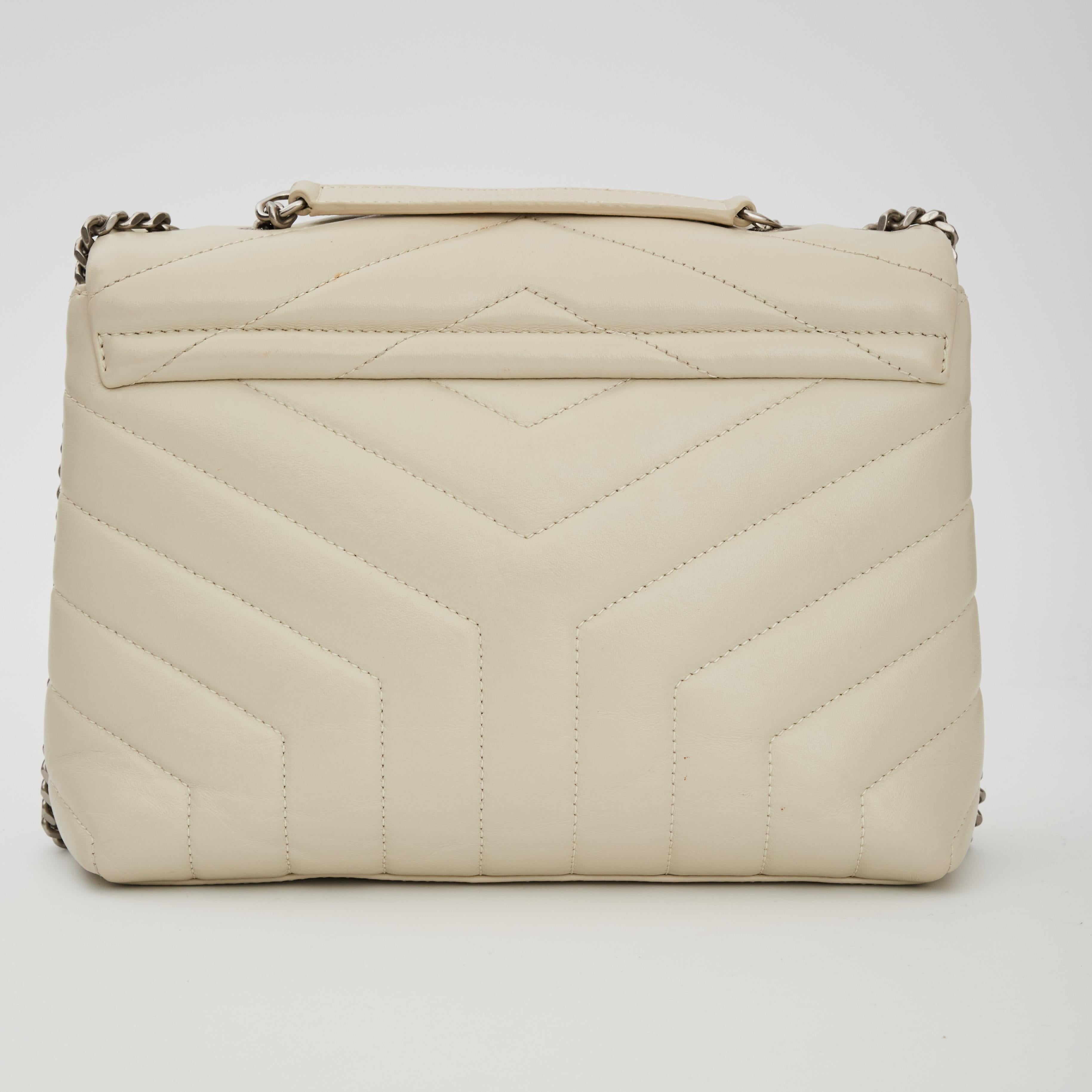 This shoulder bag features chevron quilting at both the front and the back with smooth calfskin leather in beige. The bag features a gold chain link shoulder strap with matching leather shoulder pads. The frontal flap features a prominent gold tone