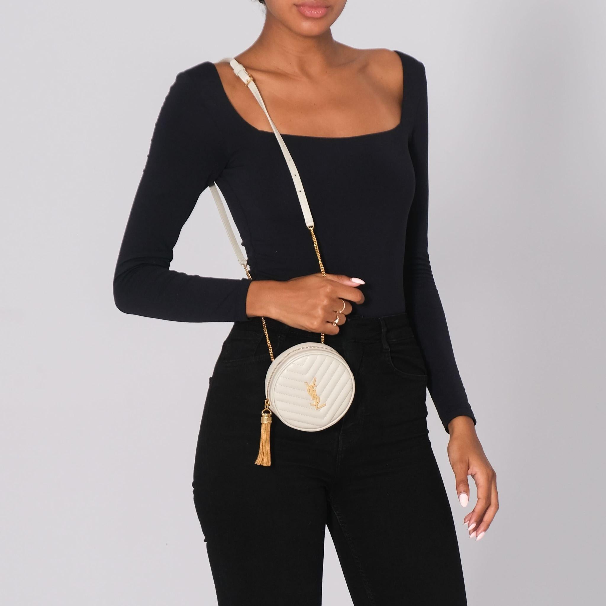 This circular crossbody is made of chevron-quilted leather in ivory. The bag features an aged gold chain-link shoulder strap, a matching top zipper, and a facing YSL monogram logo. The zipper opens to a black fabric interior with a patch pocket.
