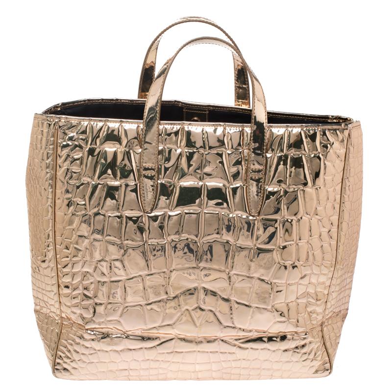 This Saint Laurent tote ensures that you carry your everyday essentials in a luxurious way. Crafted from croc-embossed patent leather in a metallic gold shade, it is provided with gold-tone hardware. It features two flat handles and studs to secure