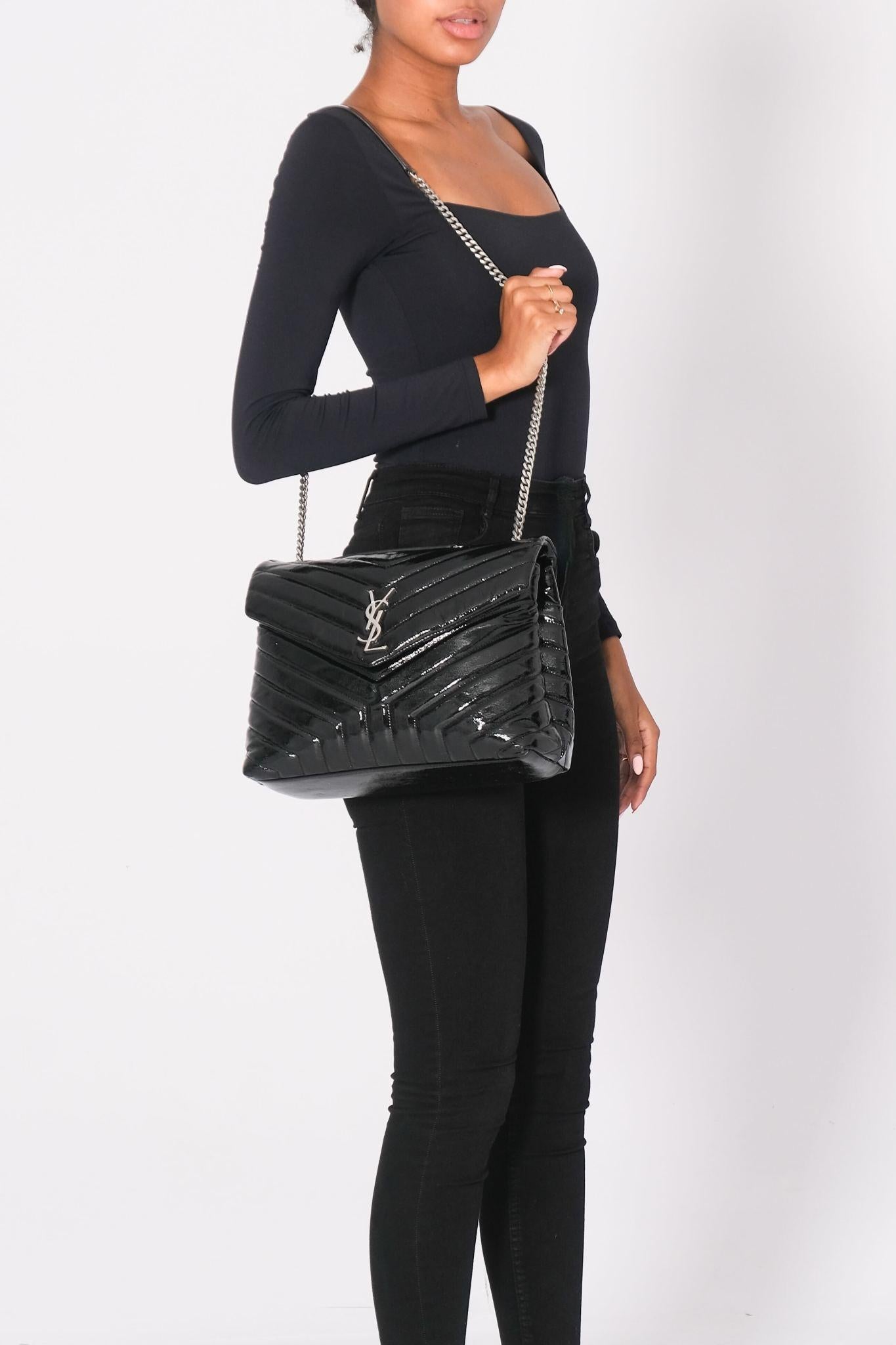 This shoulder bag is made of chevron-quilted patent leather in black. The bag features a silver chain-link shoulder strap with matching leather shoulder pads. The flap has a prominent silver YSL monogram detail and opens to a partitioned black