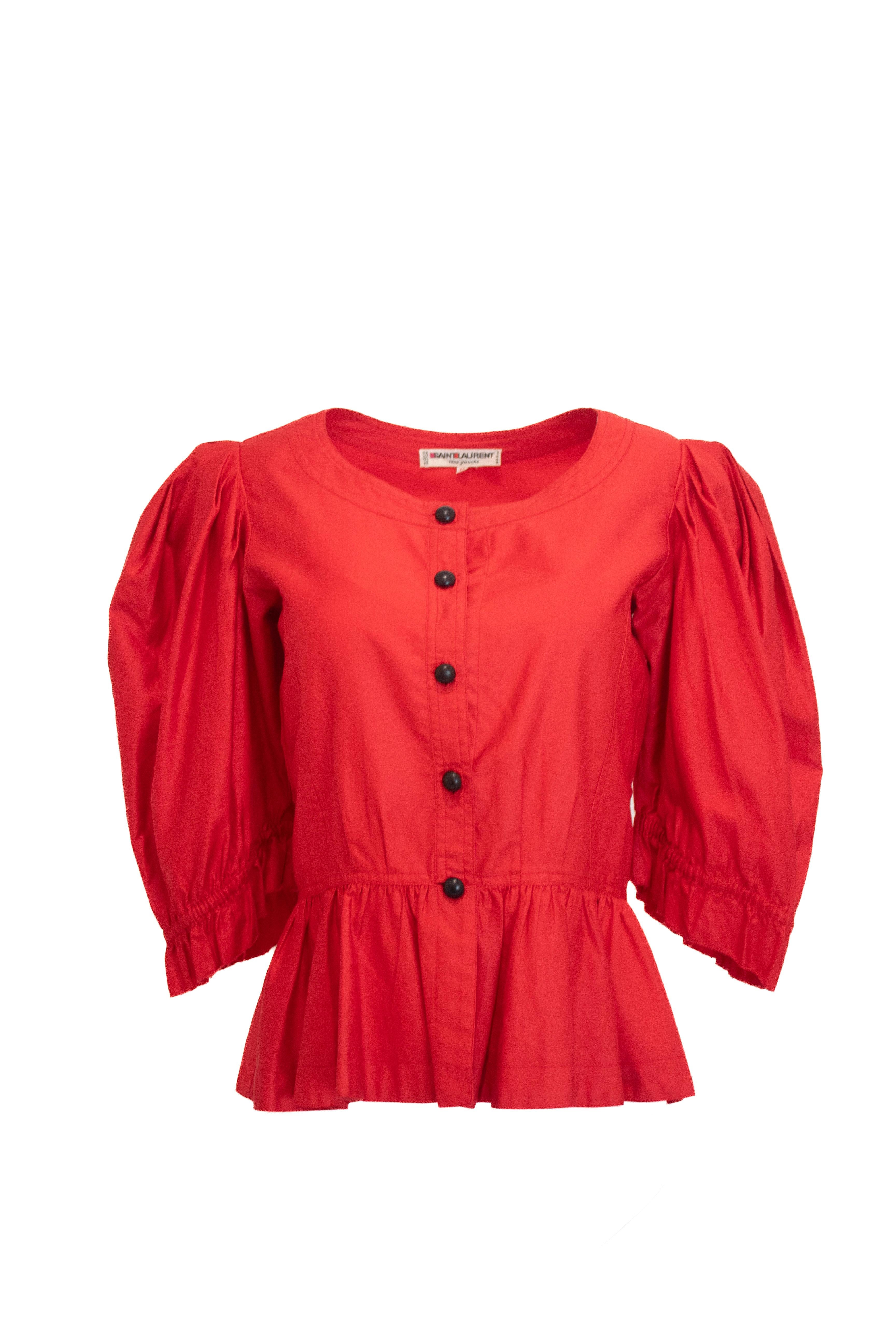 Questa blusa vintage di Saint Laurent Rive Gauche è caratterizzata da una vibrante tonalità di rosso, maniche a sbuffo e una lusinghiera silhouette a peplo accentuata da classici bottoni neri. Il suo design senza tempo lo rende un capo ideale per