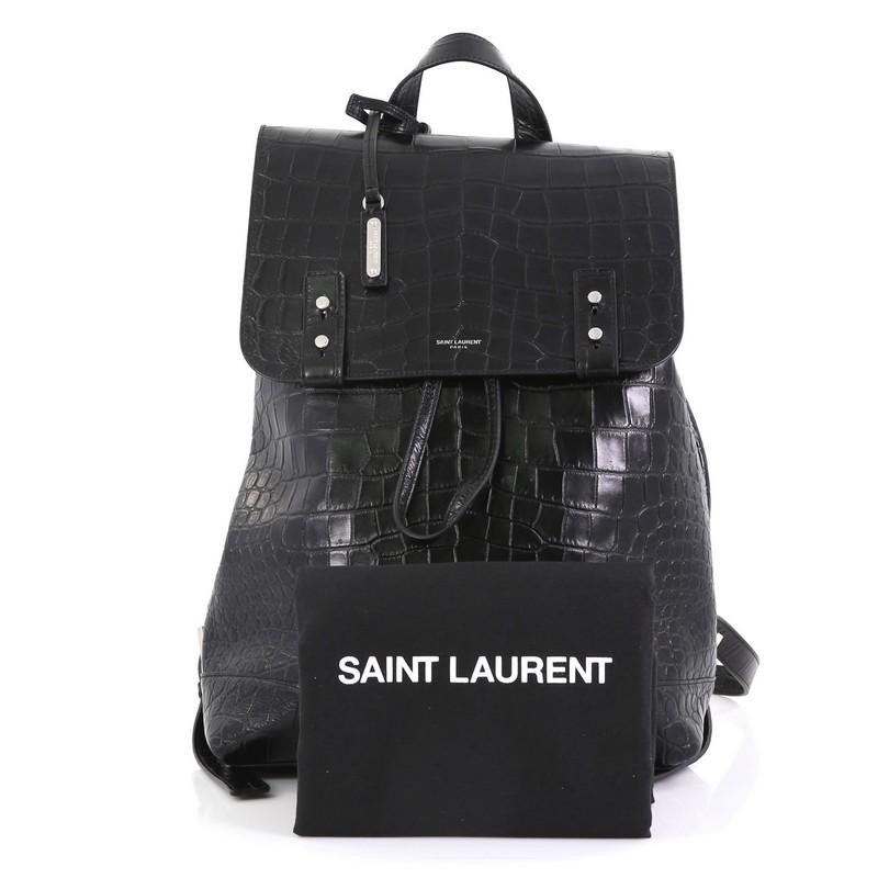 This Saint Laurent Sac de Jour Backpack Crocodile Embossed Leather Medium, crafted in black crocodile embossed leather, features adjustable leather shoulder straps, front flap and silver-tone hardware. Its drawstring closure opens to a black fabric