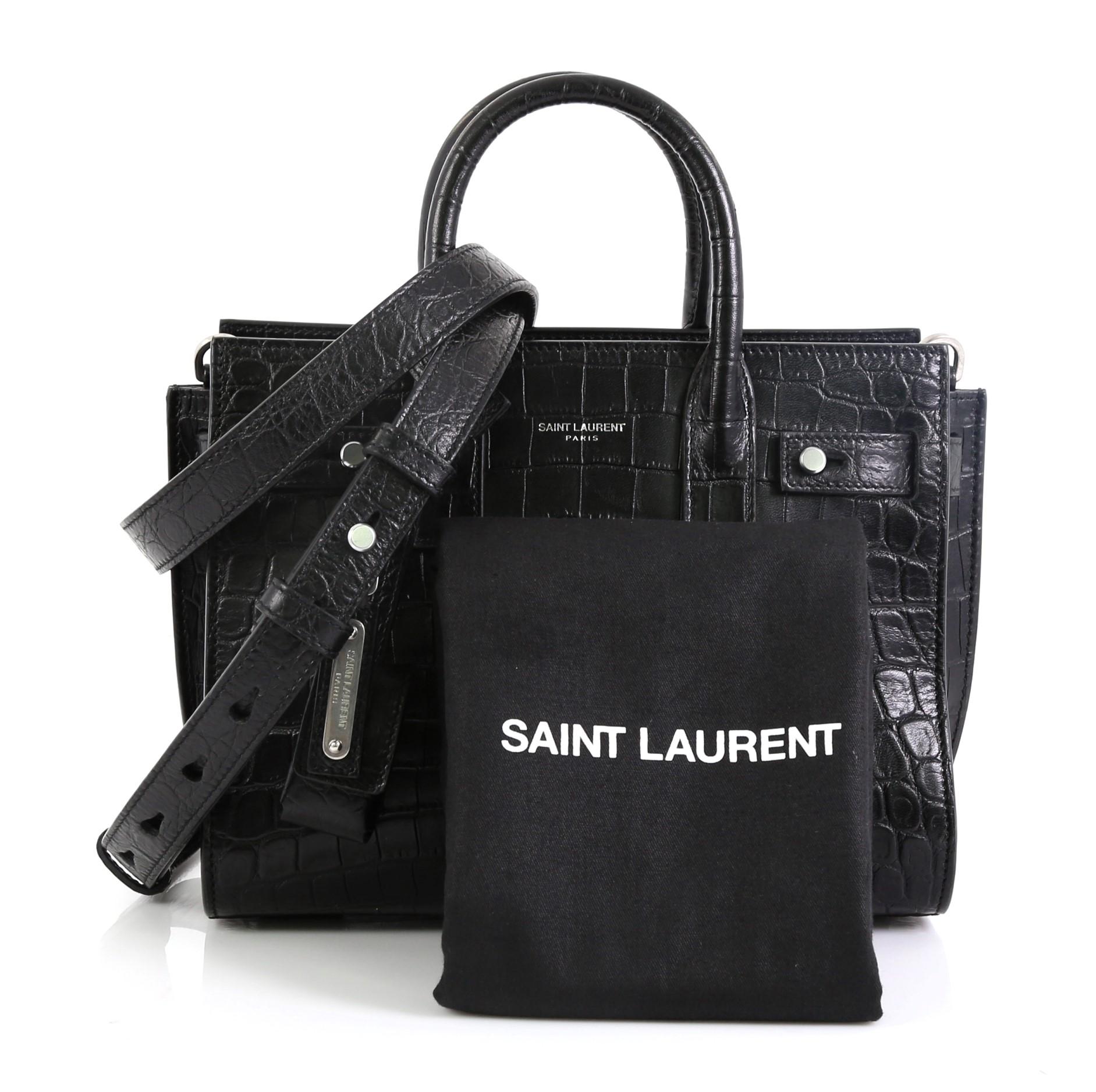 This Saint Laurent Sac de Jour Souple Bag Crocodile Embossed Leather Nano, crafted from black crocodile embossed leather, features dual rolled leather handles, accordion sides, leather tabs threaded through side gusset, and silver-tone hardware. It
