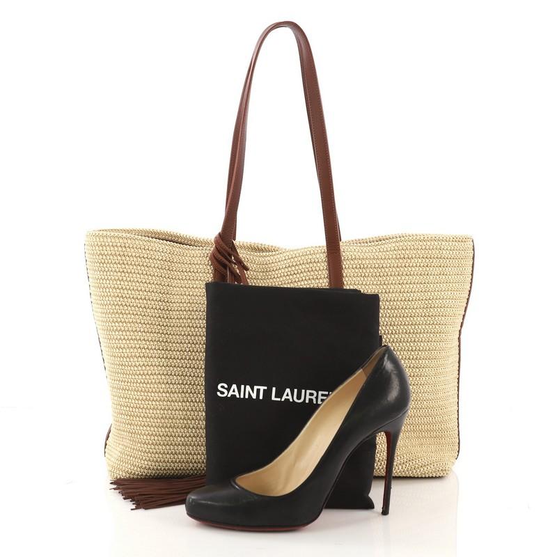This Saint Laurent Shopper Tote Woven Raffia East West, crafted from beige woven raffia, features dual flat leather handles, and gold-tone hardware. Its magnetic snap closure opens to a brown fabric interior. **Note: Shoe photographed is used as a