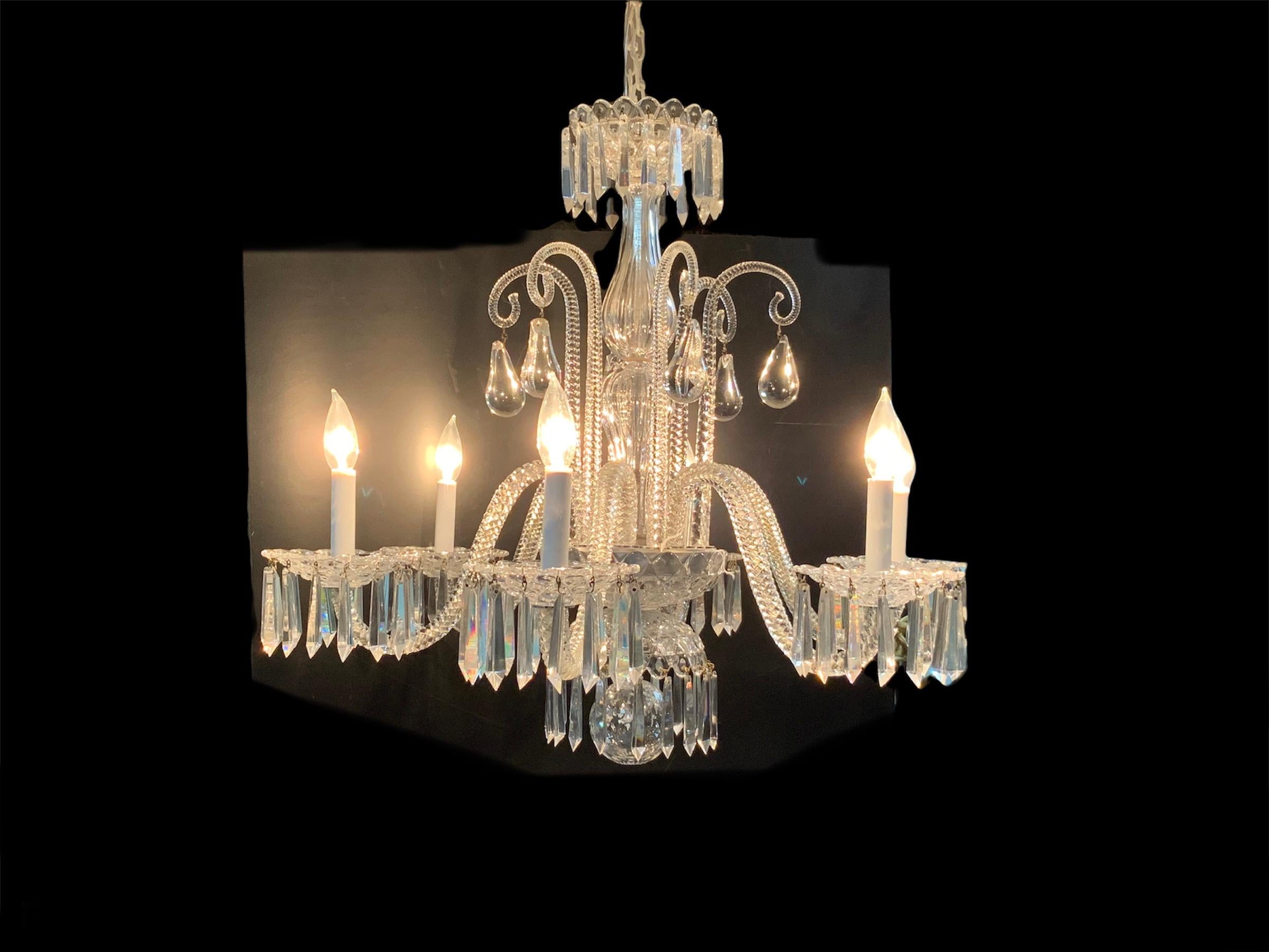 This is a cut crystal chandelier of six arms. Starting at the top, it depicts a canopy with multiple embossed hexagons shaped cut. From there come down a column formed by two fluted crystal urns that ends with a bowl with the same pattern of the