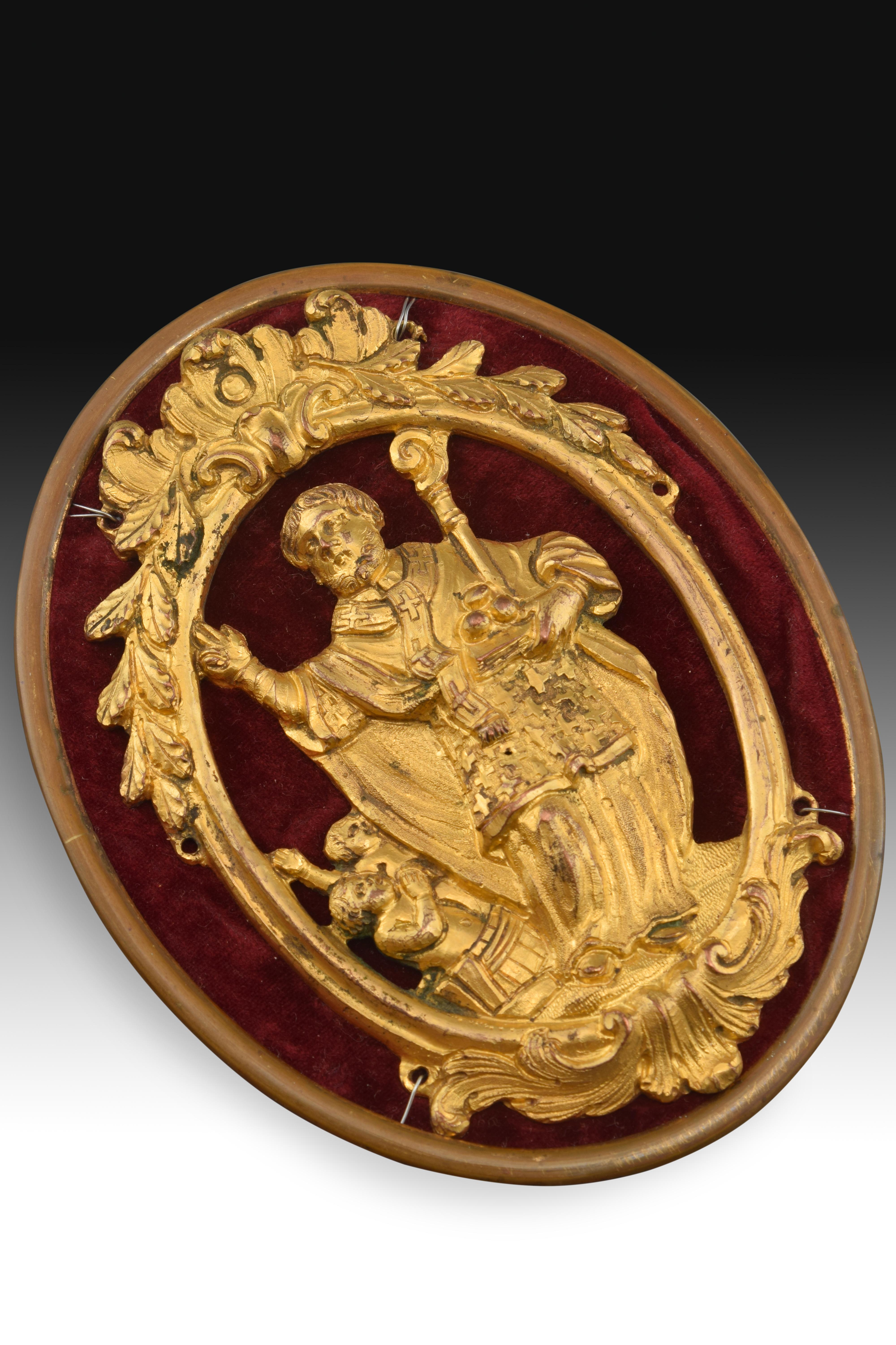 Saint Nicholas devotional medal on support. Gilded bronze, textile, metal, 18th century.
Golden bronze devotional medal placed on a support by wires that has an oval frame enhanced with a top with vegetal elements in symmetrical composition and