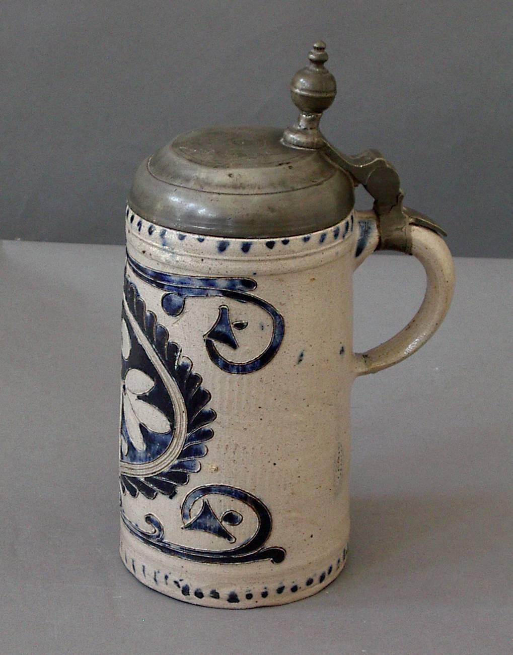 German stoneware tankard decorated with incised cartouche and swags, filled with cobalt glaze. Bottom shows the coil construction. Westerwald, 18th century. Pewter cover with finial-shaped thumb latch.