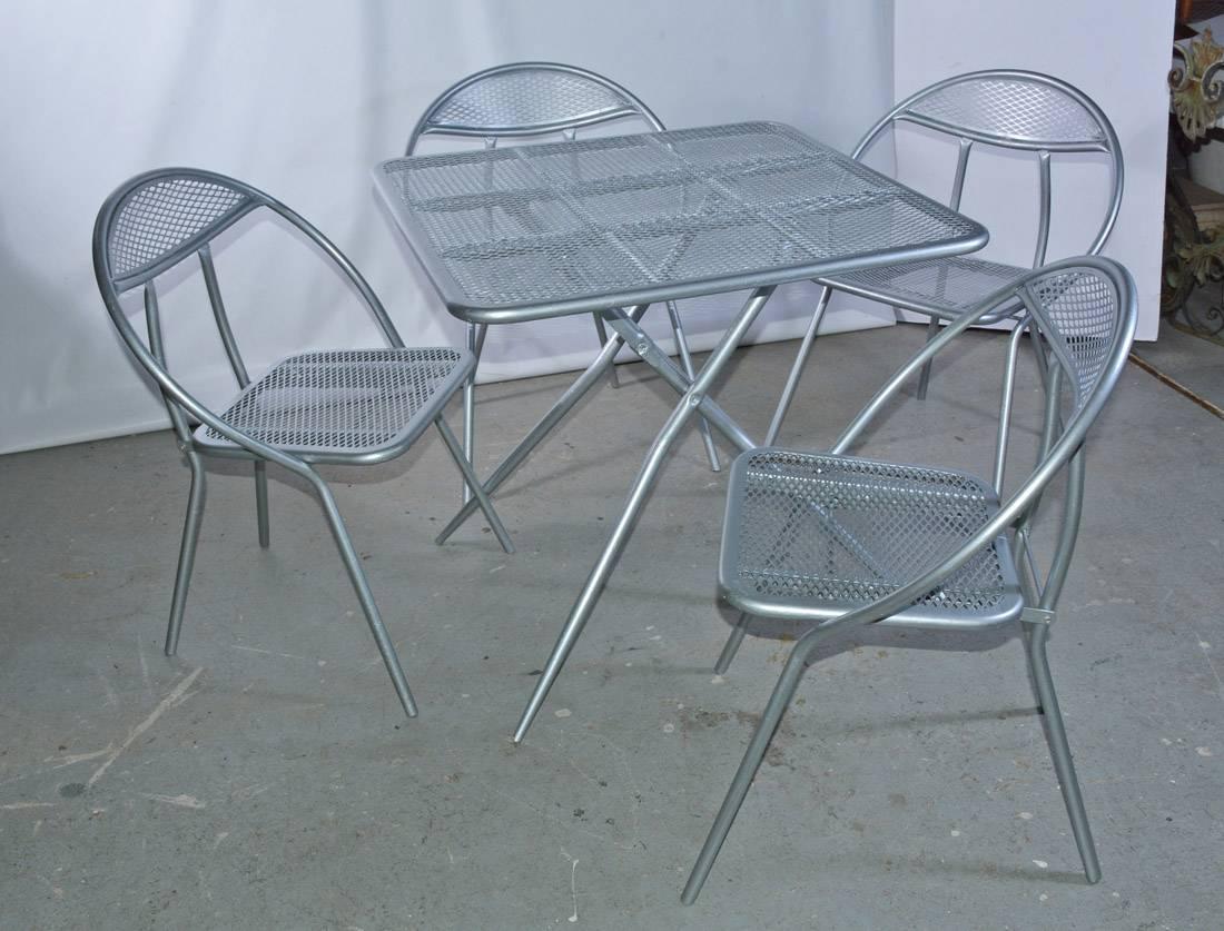 The Mid-Century Modern metal patio or garden table and matching four chair dining set, painted silver, fold flat for storage purposes. The square table has a mesh surface, while the chairs with rounded-back have mesh seats and partially mesh backs.