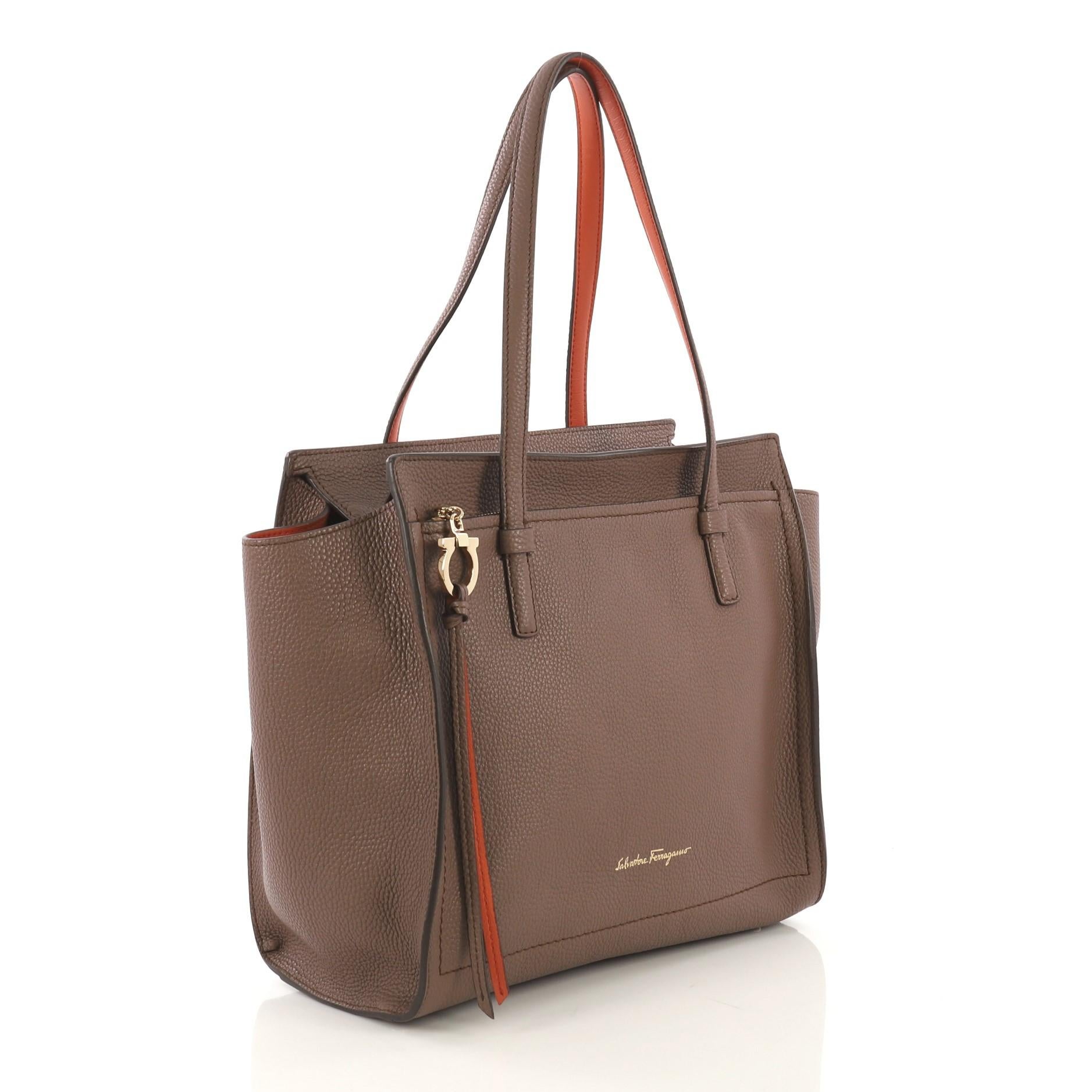 This Salvatore Ferragamo Amy Tote Pebbled Leather Medium, crafted from taupe pebbled leather, features exterior front zip pocket with Gancio charm zip pull, protective base studs and gold-tone hardware. Its zip closure opens to an orange leather