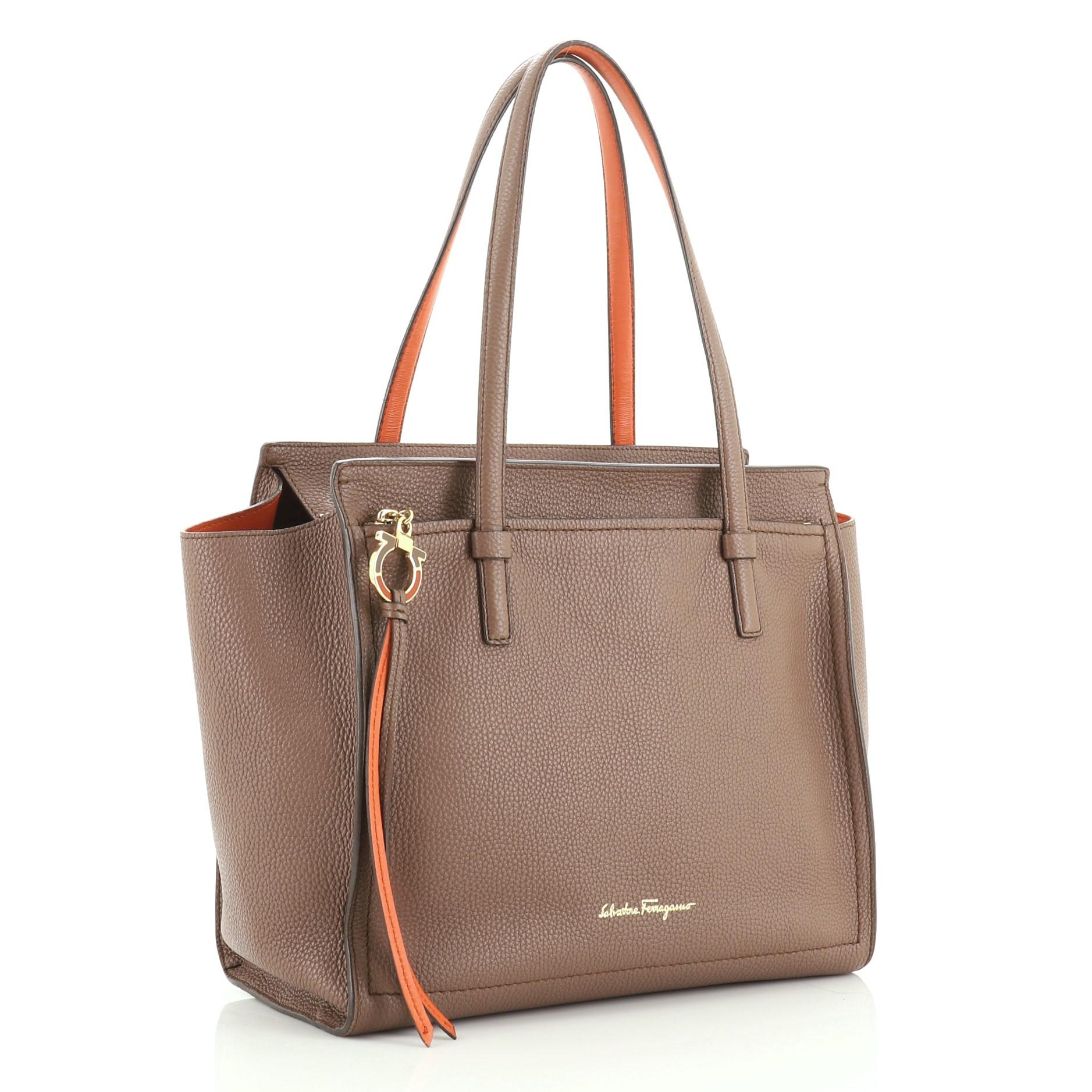 This Salvatore Ferragamo Amy Tote Pebbled Leather Medium, crafted from neutral pebbled leather, features dual flat shoulder strap, exterior front zip pocket with Gancio zip pull, protective base studs and gold-tone hardware. Its zip closure opens to