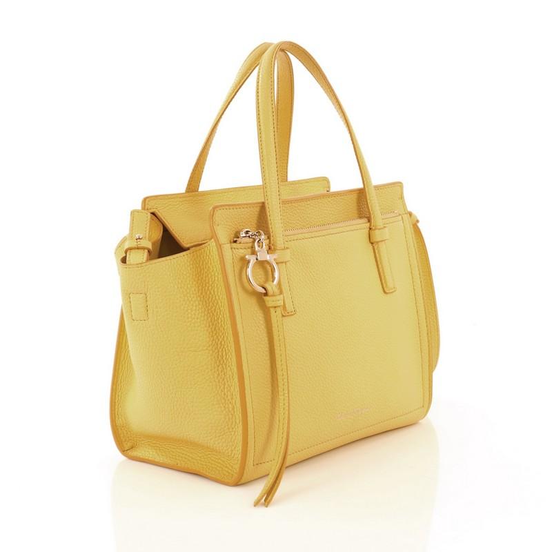 This Salvatore Ferragamo Amy Tote Pebbled Leather Mini, crafted from yellow pebbled leather, features dual flat handles, exterior front zip pocket with Gancio charm zip pull, Salvatore Ferragamo logo lettering at bottom center, protective base studs