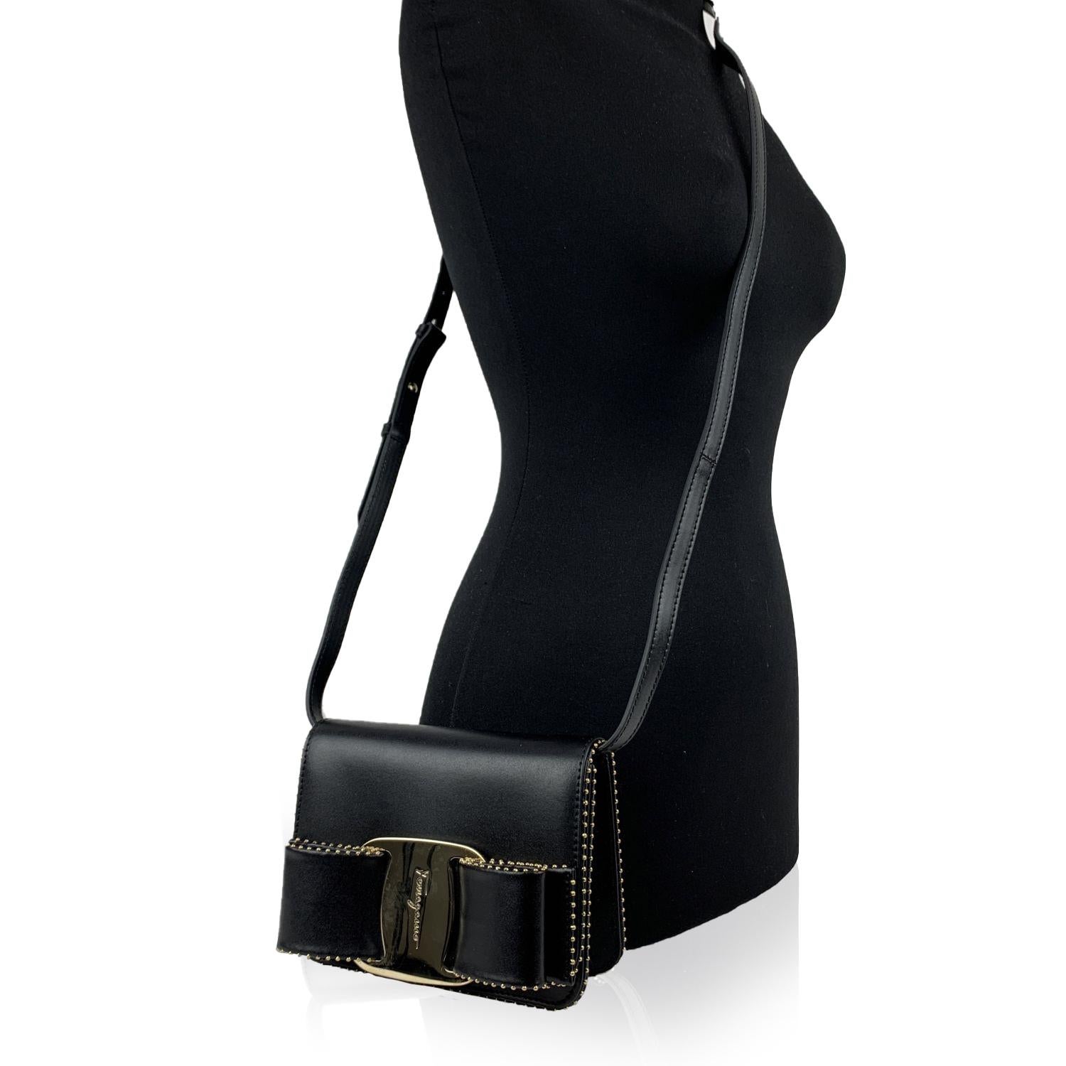 Beautiful 'Vara RW O' small shoulder bag by Salvatore Ferragamo. Made of black calf leather. It features a flap closure with iconic Vara Bow detailing on the front and gold metal micro-studs detailing around the edges of the bag. Adjustable and