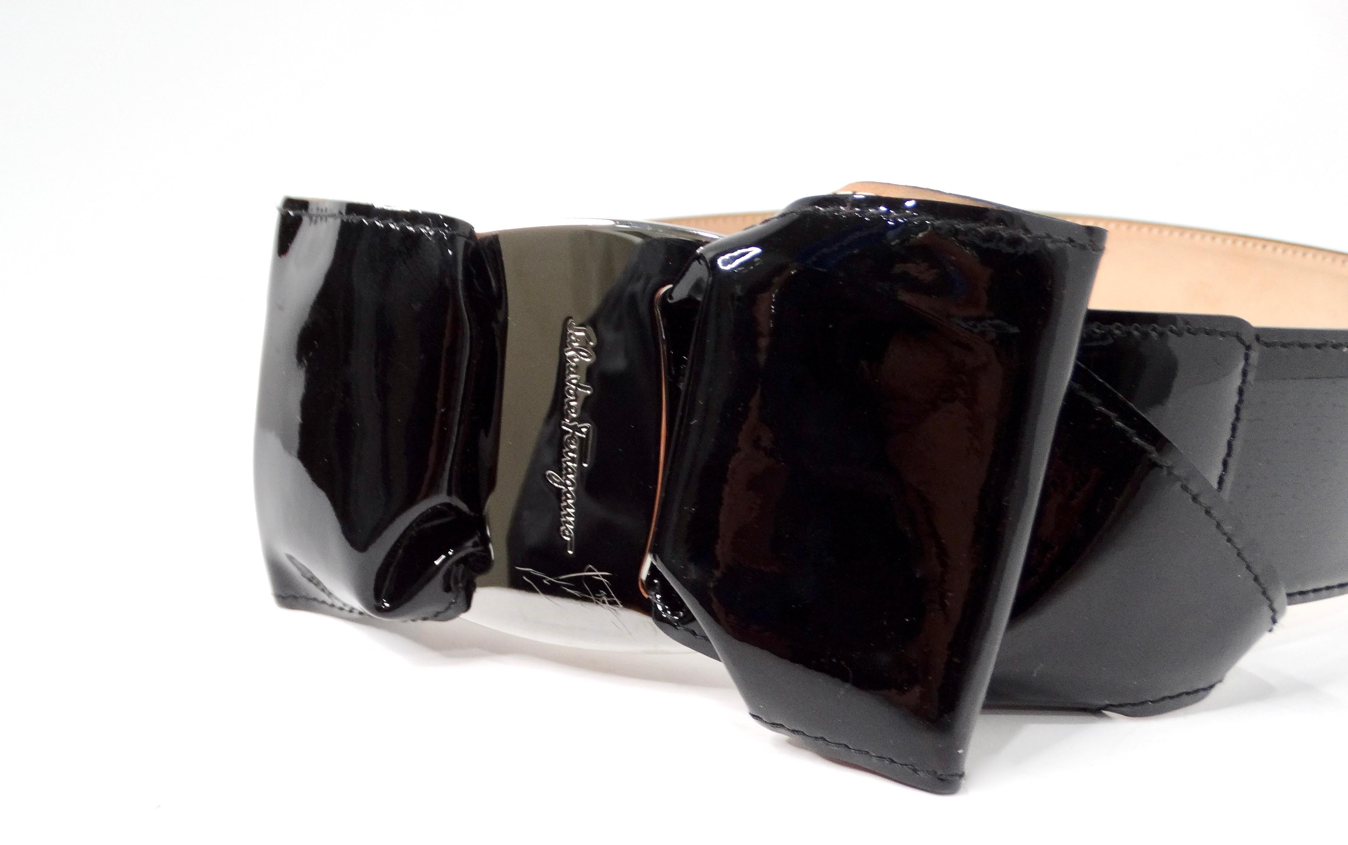 The Salvatore Ferragamo Black Patent Leather Bow Style Belt is a chic and sophisticated accessory that effortlessly elevates any outfit. Featuring a stunning bow-shaped buckle adorned with a silver-tone metal centerpiece and embossed logo, this belt