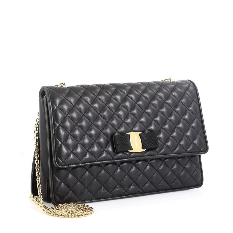 This Salvatore Ferragamo Ginny Crossbody Bag Quilted Leather Medium, crafted from black quilted leather, features the Vara bow design with Ferragamo logo plate, chain link strap, and gold-tone hardware. Its hidden magnetic snap closure opens to a