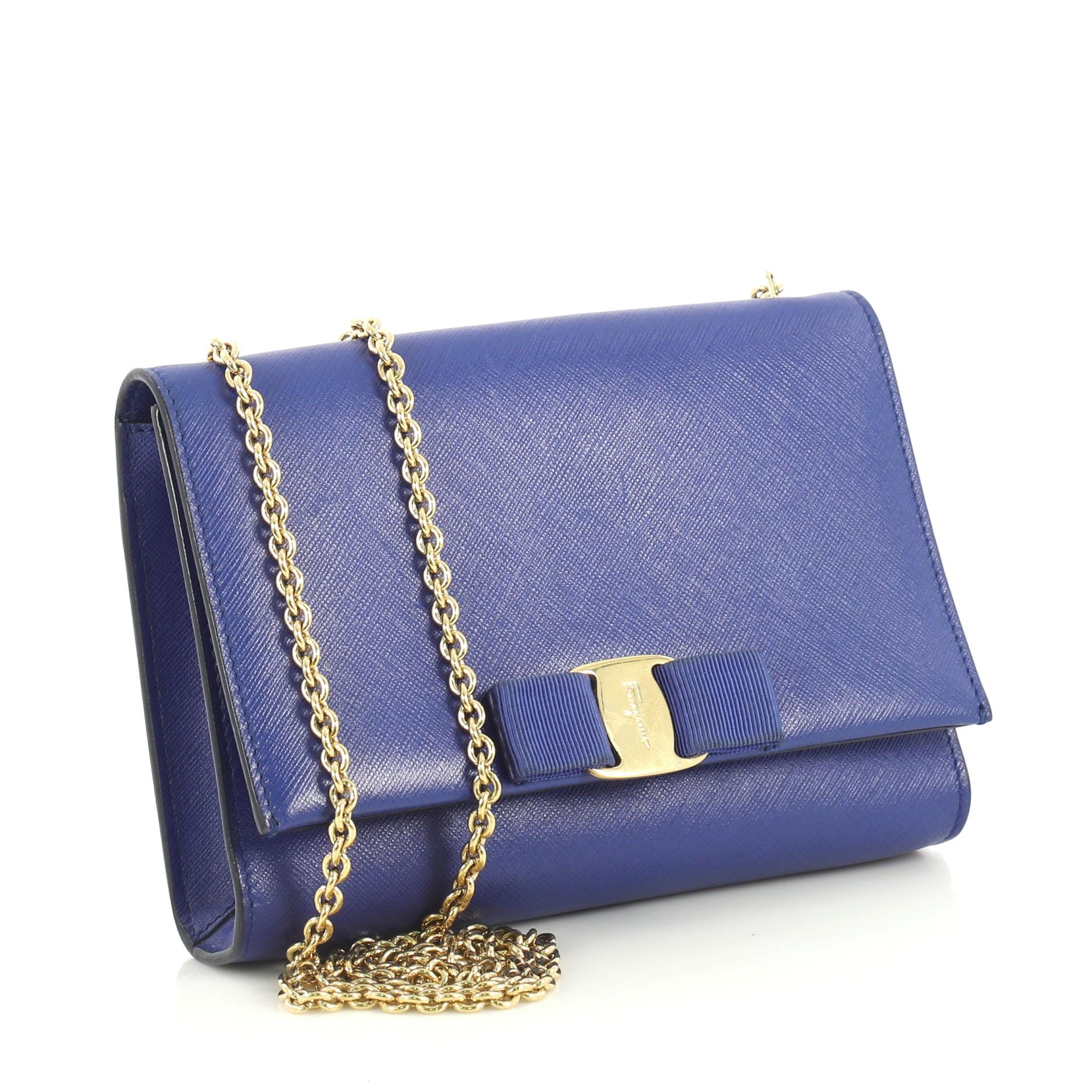 This Salvatore Ferragamo Ginny Crossbody Bag Saffiano Leather Small, crafted from blue saffiano leather, features chain link strap, Vara bow design with Ferragamo logo plate, and gold-tone hardware. Its hidden magnetic snap closure opens to a blue