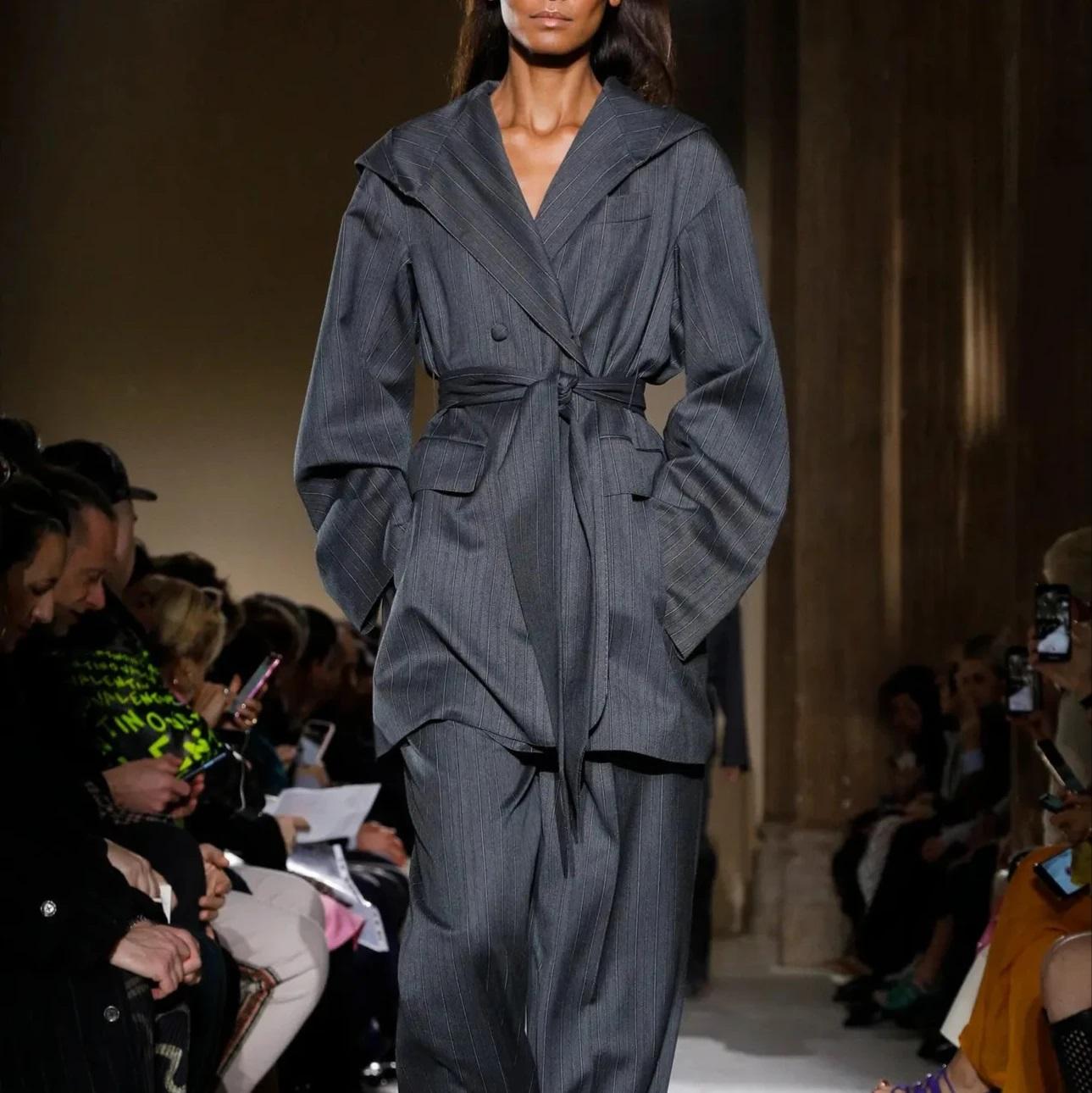 Abito gessato in lana vergine grigia dalla passerella Autunno 2019. Presenta una giacca con cappuccio con cintura e pantaloni a vita alta a pieghe a gamba larga per una silhouette audace e raffinata.
 Osservazioni: ci sono due buchi sui pantaloni. 
