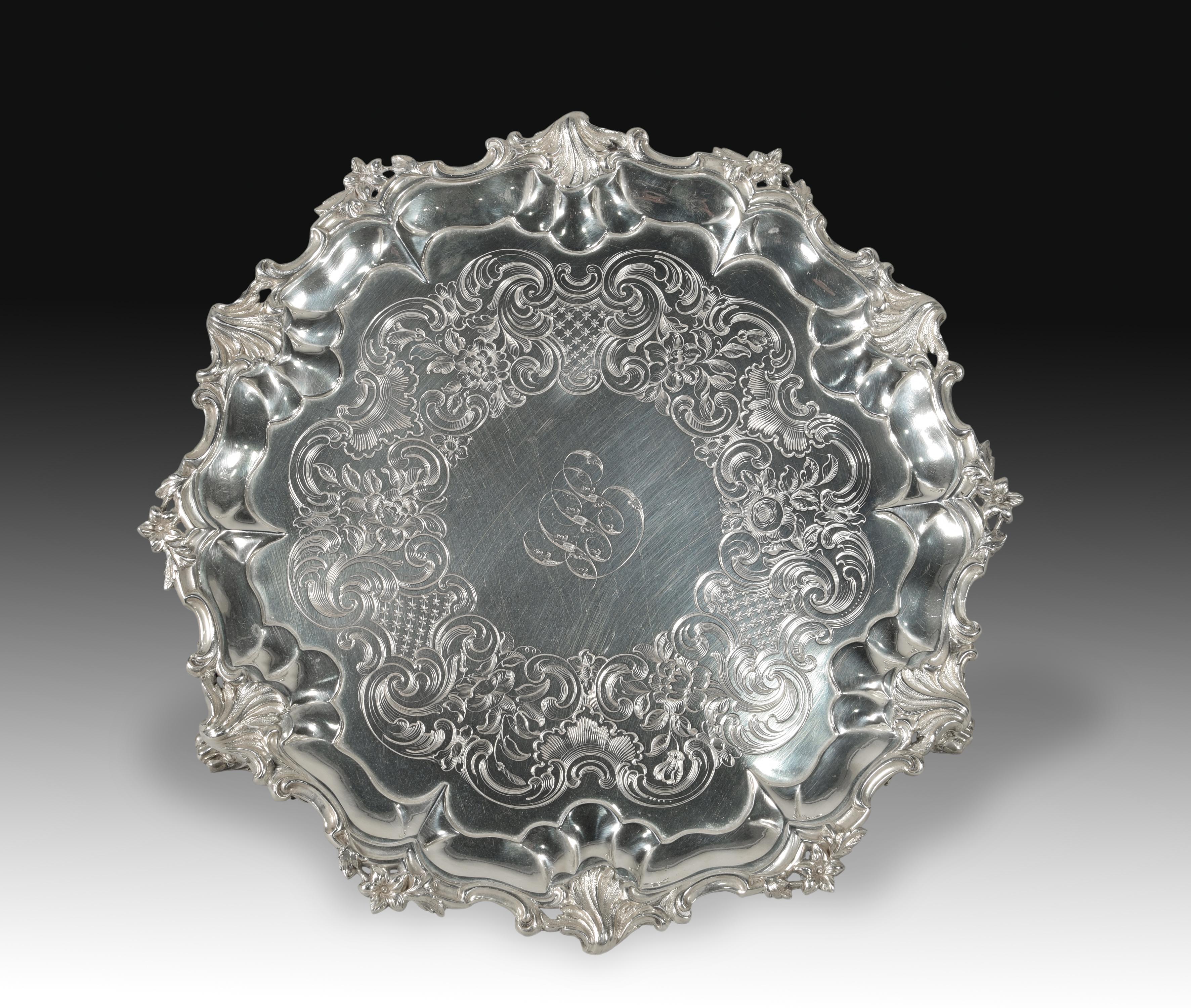Salver. Silver. Edward, John & William Barnard. England, London, 1862. With contrast markings.
Round salvilla with mixed contours decorated with elements in slight relief towards the edge and engravings towards the center, arranged around initials,