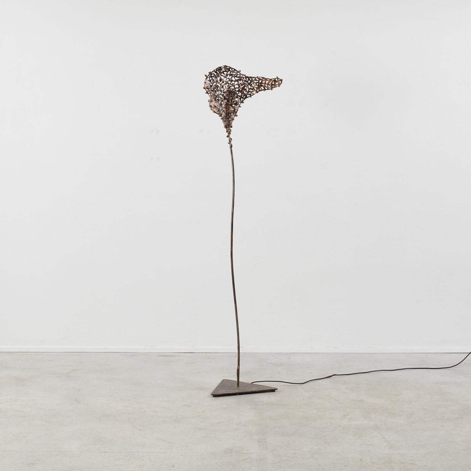 Una base triangolare si innalza in uno stelo sottile e dolcemente curvo, che culmina in una sorgente luminosa racchiusa da un reticolo scultoreo di frammenti di ferro. La lampada da terra Nuova Arte proietta e interrompe la luce in egual misura,