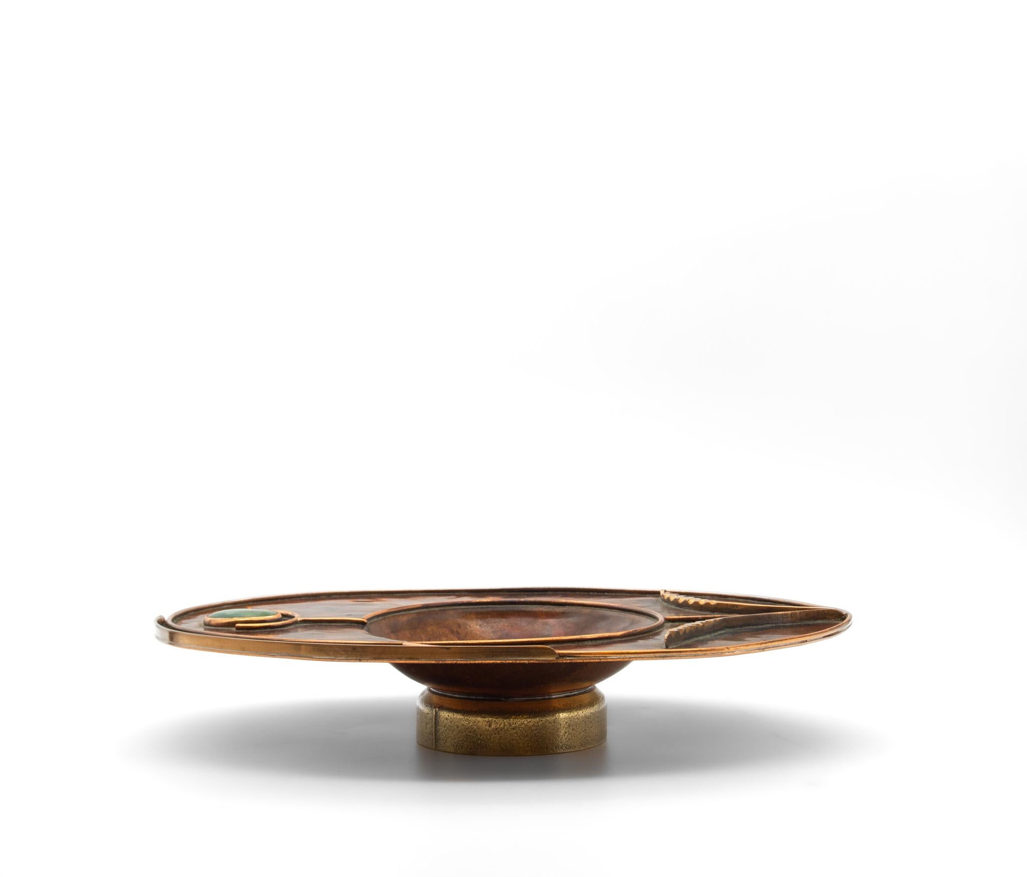 Un piatto stilizzato in rame di Sussex Guild, con un disegno di grano strombato e un inserto di pietra verde cabochon su una base in ottone. Di Sam Fanaroff (1926-2019). Iniziali timbrate dall'artista. 

Questo piatto decorativo è in ottime