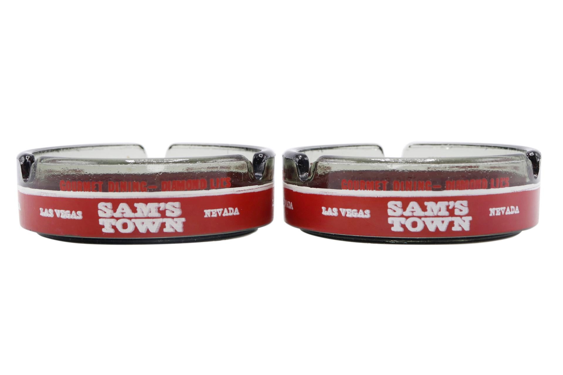 A pair of gray glass ashtrays branded Sam's Town, Las Vegas, Nevada. The bases are decorated with concentric circles and the outside with red lettering and white lettering on a white background. Sam's Town Hotel and Gambling Hall, abbreviated to