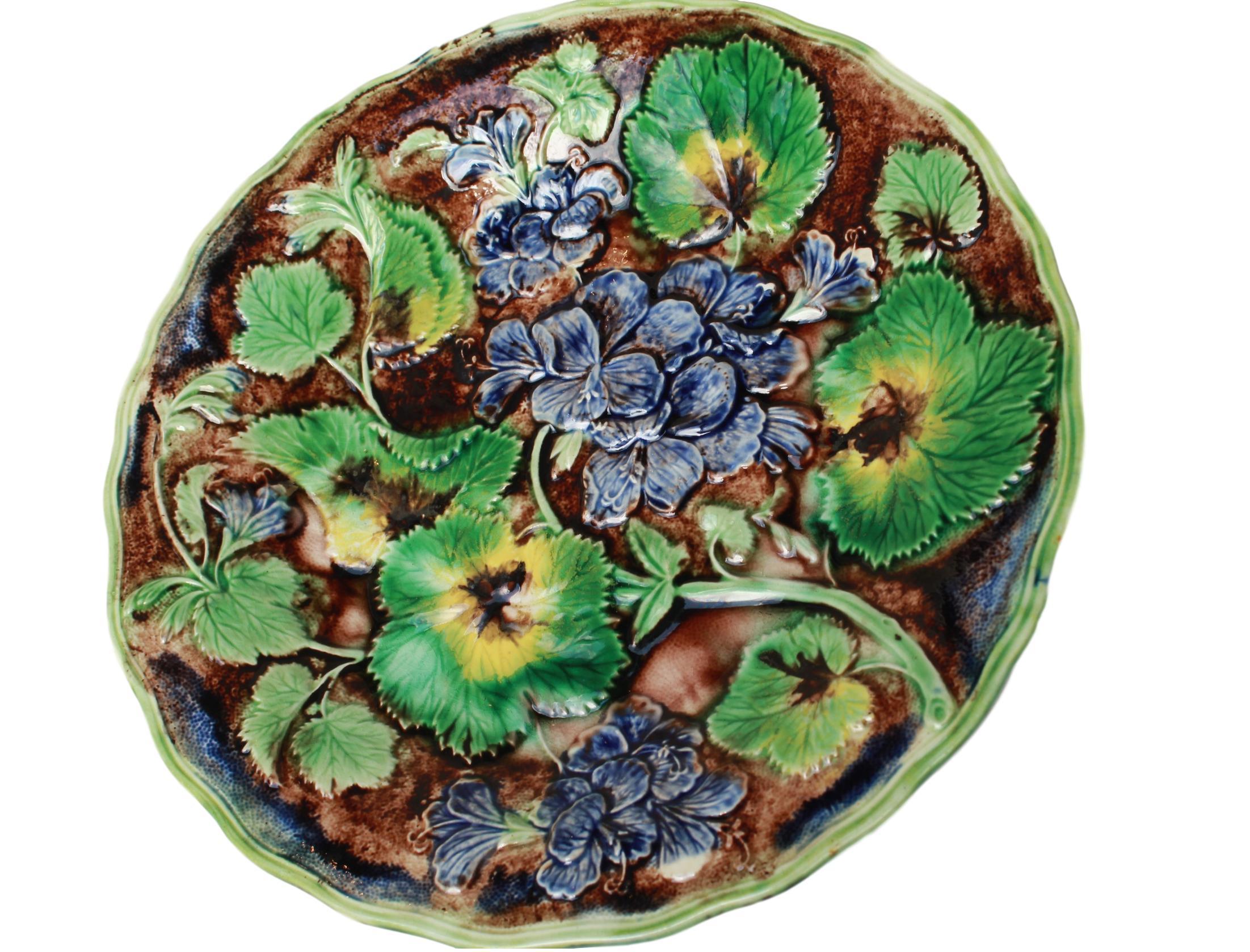 Samuel Alcock & Co. Majolica purple glazed geraniums plate, English, circa 1860, molded with purple glazed geranium blossoms, stems and leave glazed in green, yellow and brown, reserved on a faux bois ground within a shaped green glazed rim.
In