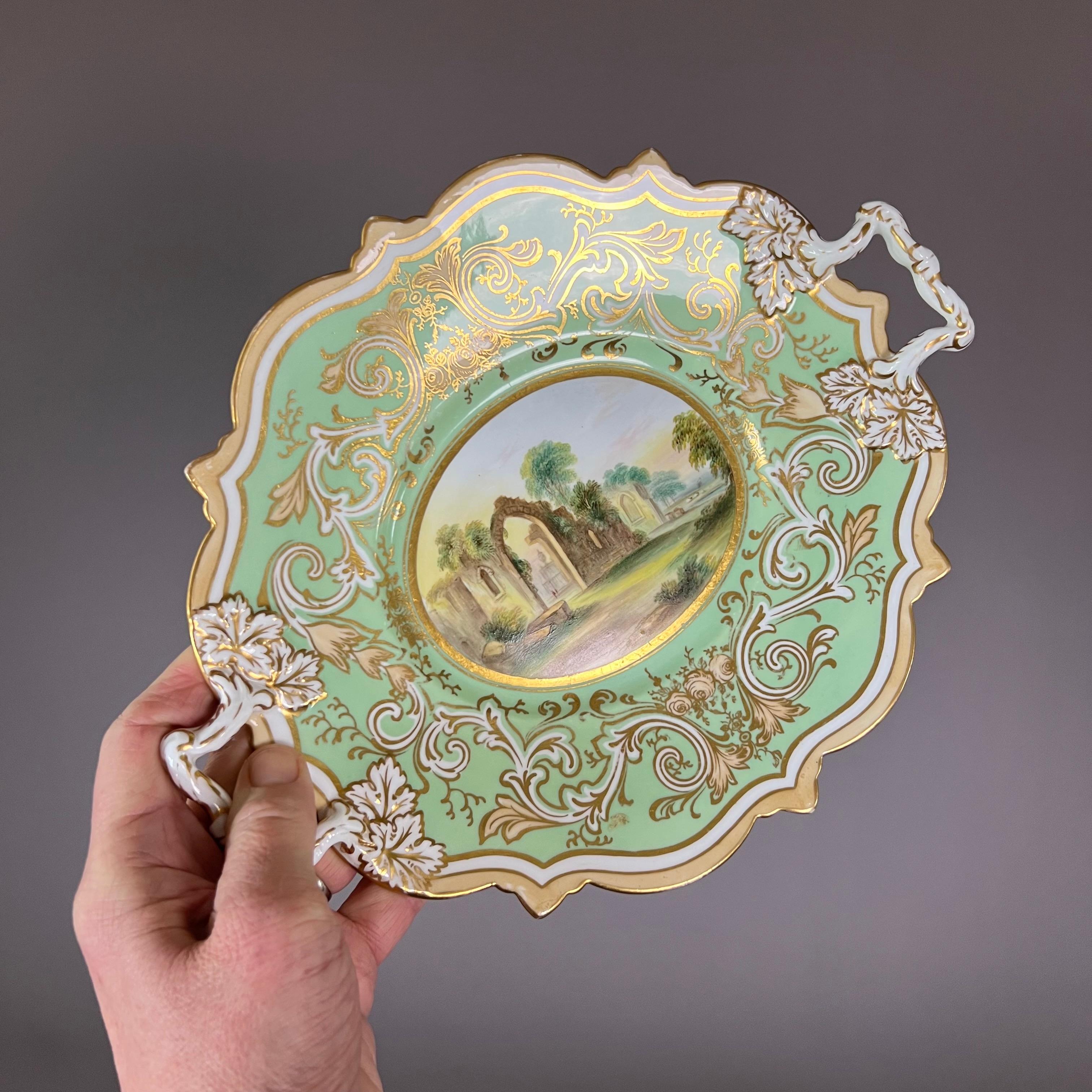 An oval low-footed comport with two handles and an octagonally scrolled shape, a moulded surface with pale yellow and white scrolling foliage on a sage green ground, and a stunning landscape in the centre of a ruin with two visitors

Pattern