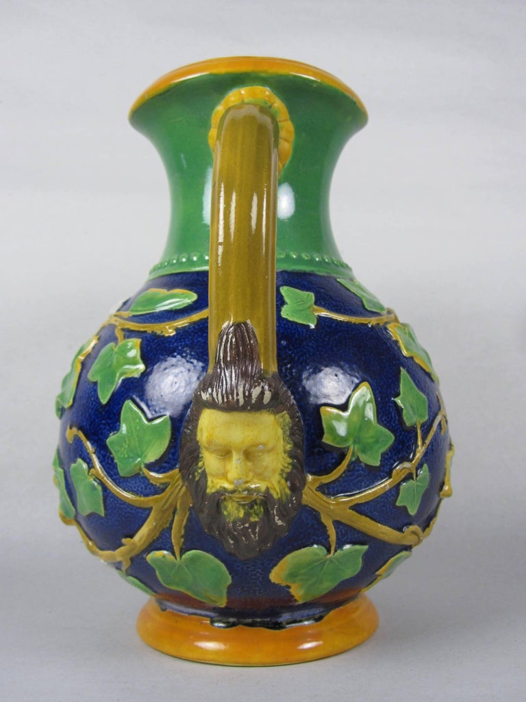 A rarely found English Majolica mask handled and climbing Ivy water pitcher, marked Samuel Alcock & Co. circa 1875. 

A round bellied cobalt blue body with a broad green collar, a yellow scrolled lip and a brown handle terminating in a mask of a