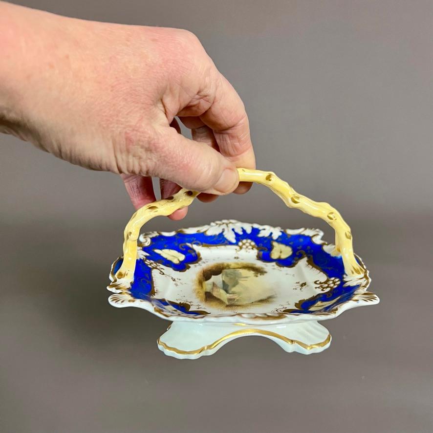 A small porcelain basket in Rococo Revival style with scroll and shell moulded borders, pierced handles on both ends, a pale yellow twig handle, a French blue ground with pale yellow foliage and a fine landscape painting in the centre.

This would