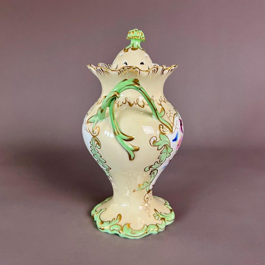 A potpourri vase with double cover, pale yellow/beige ground with apple green details, Rococo shape with fruit finial and scroll handles and foot; a flower reserve on one side and a landscape reserve on the other, both beautifully hand