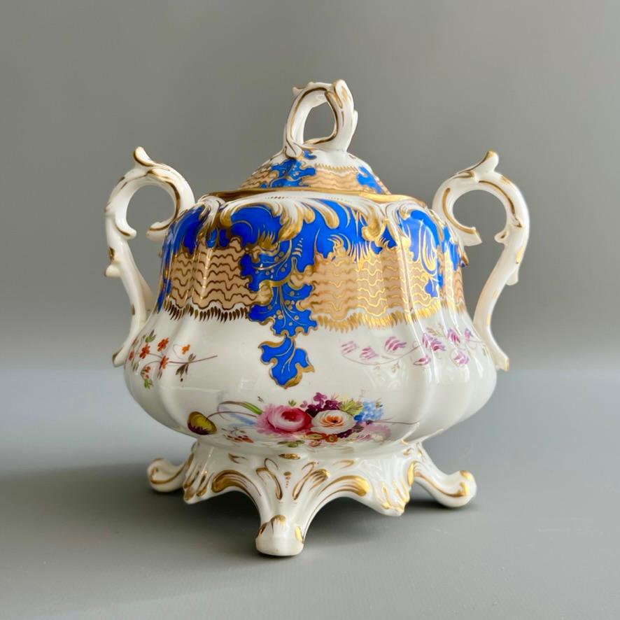 Ein birnenförmiges Rokoko-Revival-Sucrier mit Deckel, mit einem kapriziösen Muster aus französisch-blauen Schriftrollen, pfirsichfarbenen und vergoldeten Wellenkartuschen und wunderschön handgemalten Blumenranken um den Korpus.

Muster 1/813
Jahr: