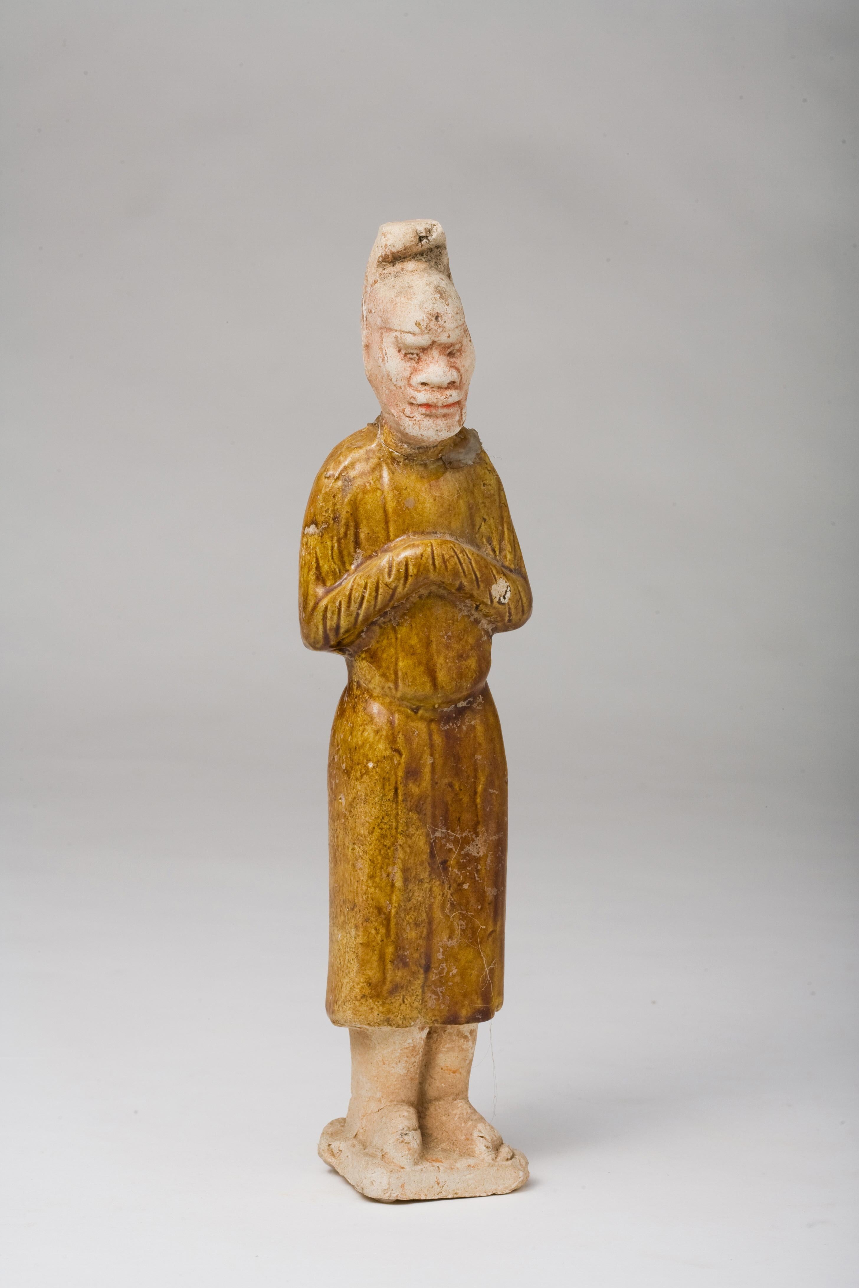 The figure stands with arms crossed in front, a pose that is often seen in tomb figurines which were intended to serve the deceased in the afterlife. The long robe and facial features of the figure are intricately crafted, showcasing the meticulous